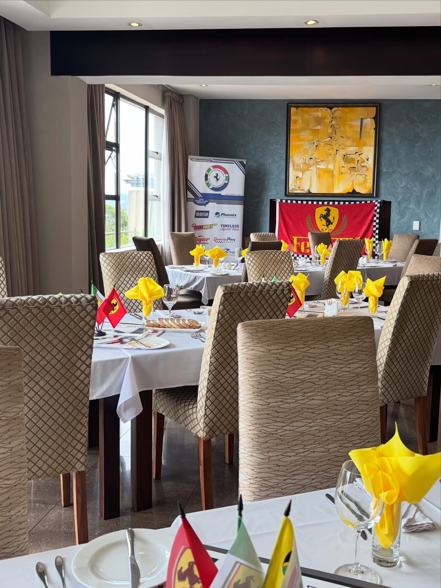 Dining tables with tan chairs, white tablecloths, and yellow napkins in a room featuring a Ferrari banner and wall art.