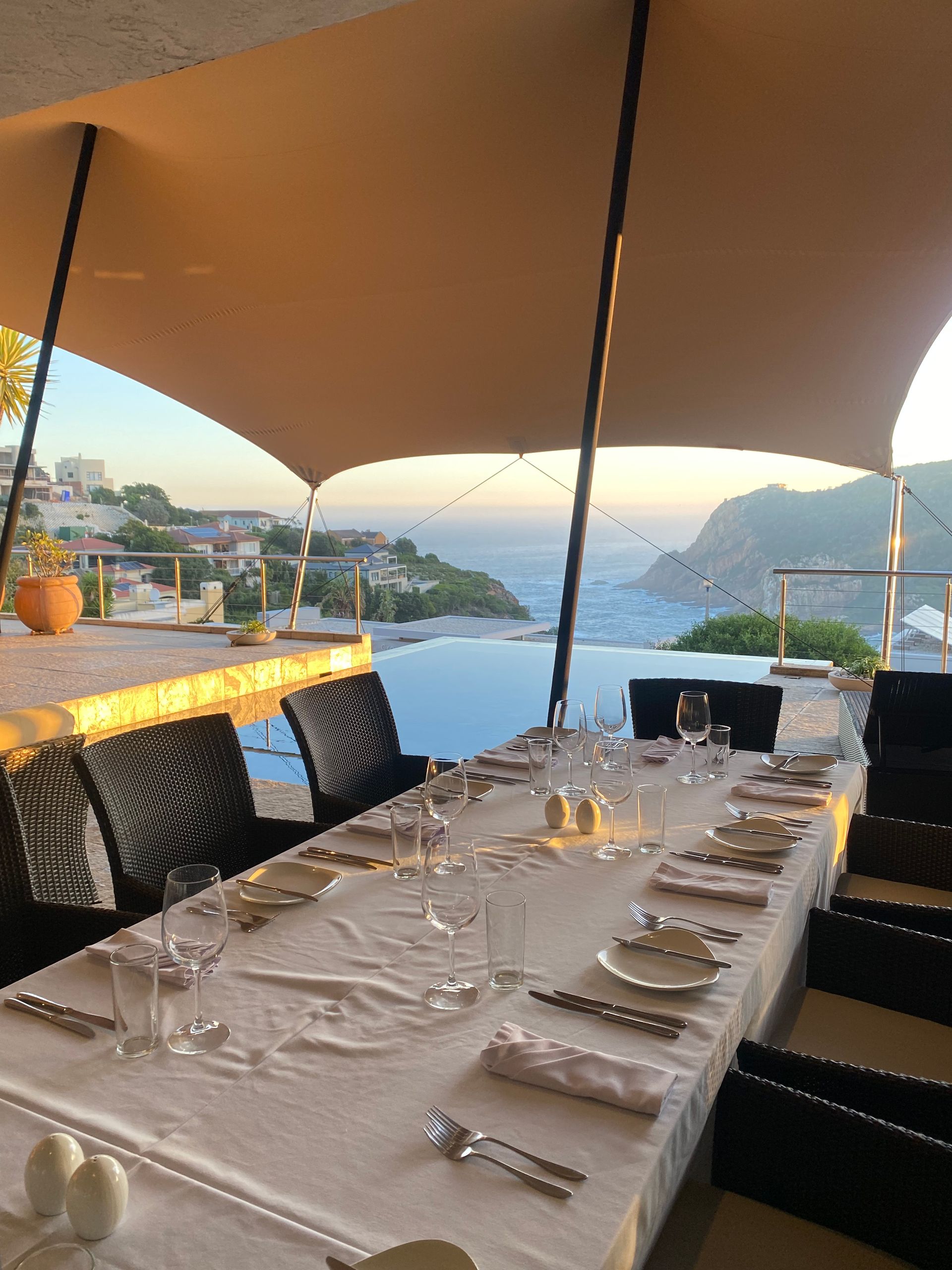 A formal dinner table set under a beige canopy with a scenic ocean and cliffside view in the background at sunset.