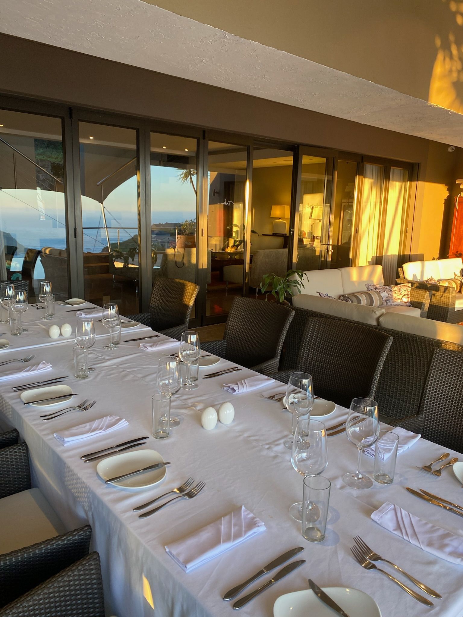 An elegantly set long dining table on a patio, overlooking a sunset view through large glass doors.