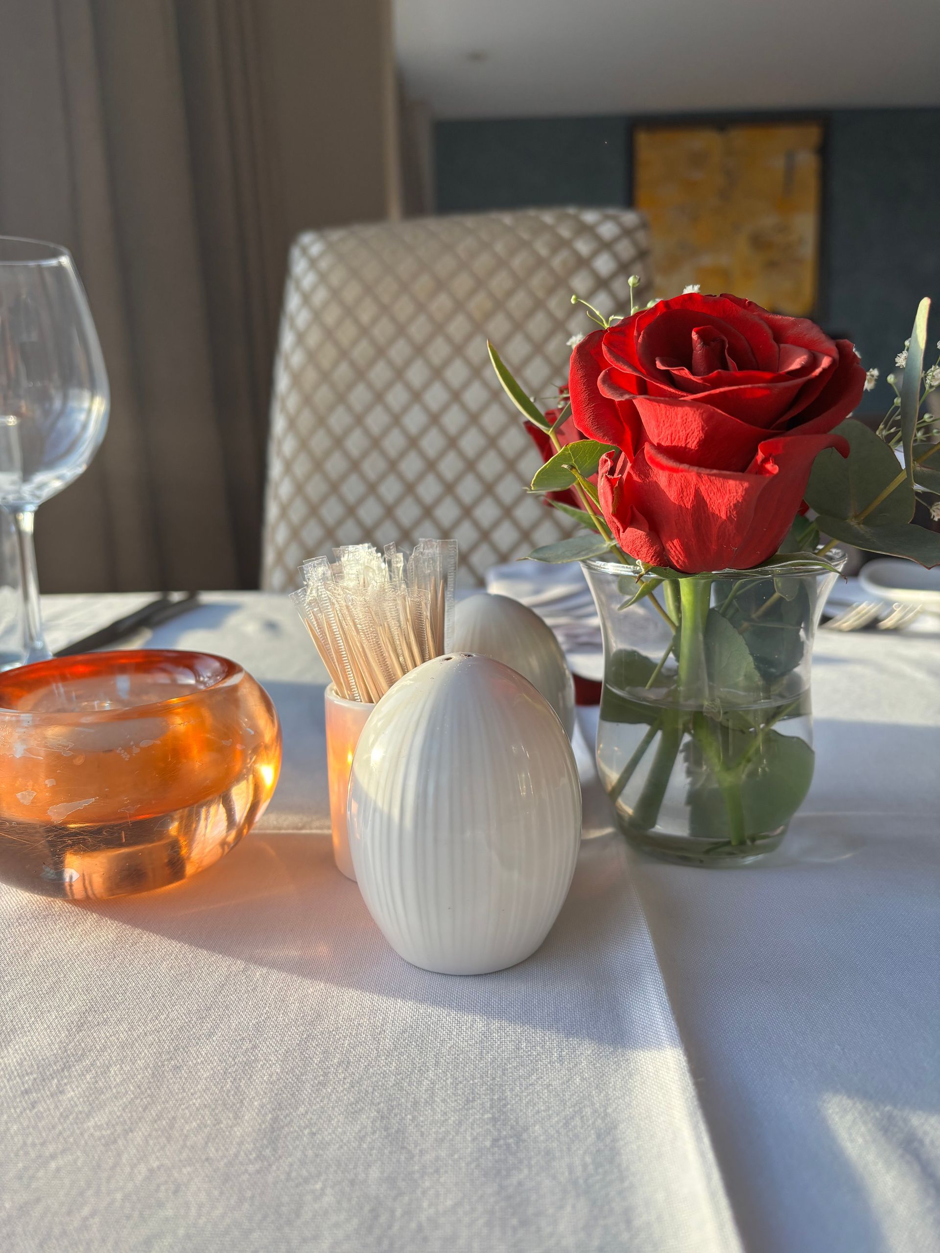 A red rose in a glass vase, two white ceramic egg-shaped shakers, and an amber glass candle holder on a white table.