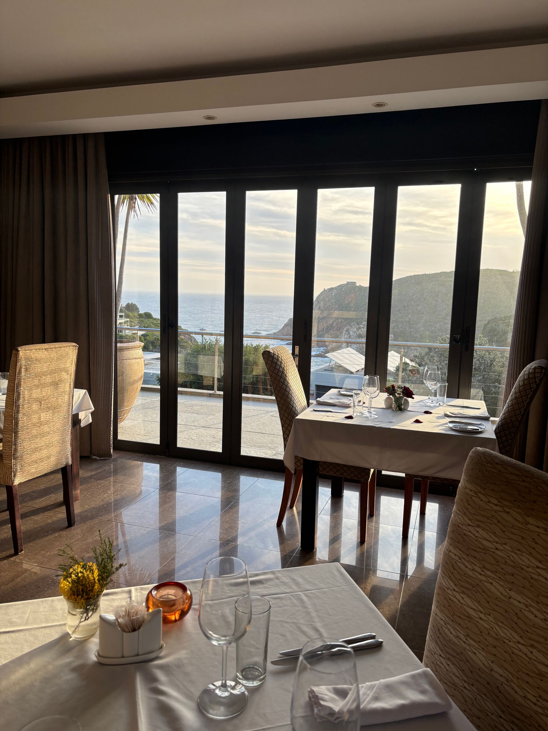 A restaurant interior with two tables set for dining, facing a wall of windows overlooking a scenic coastal view.
