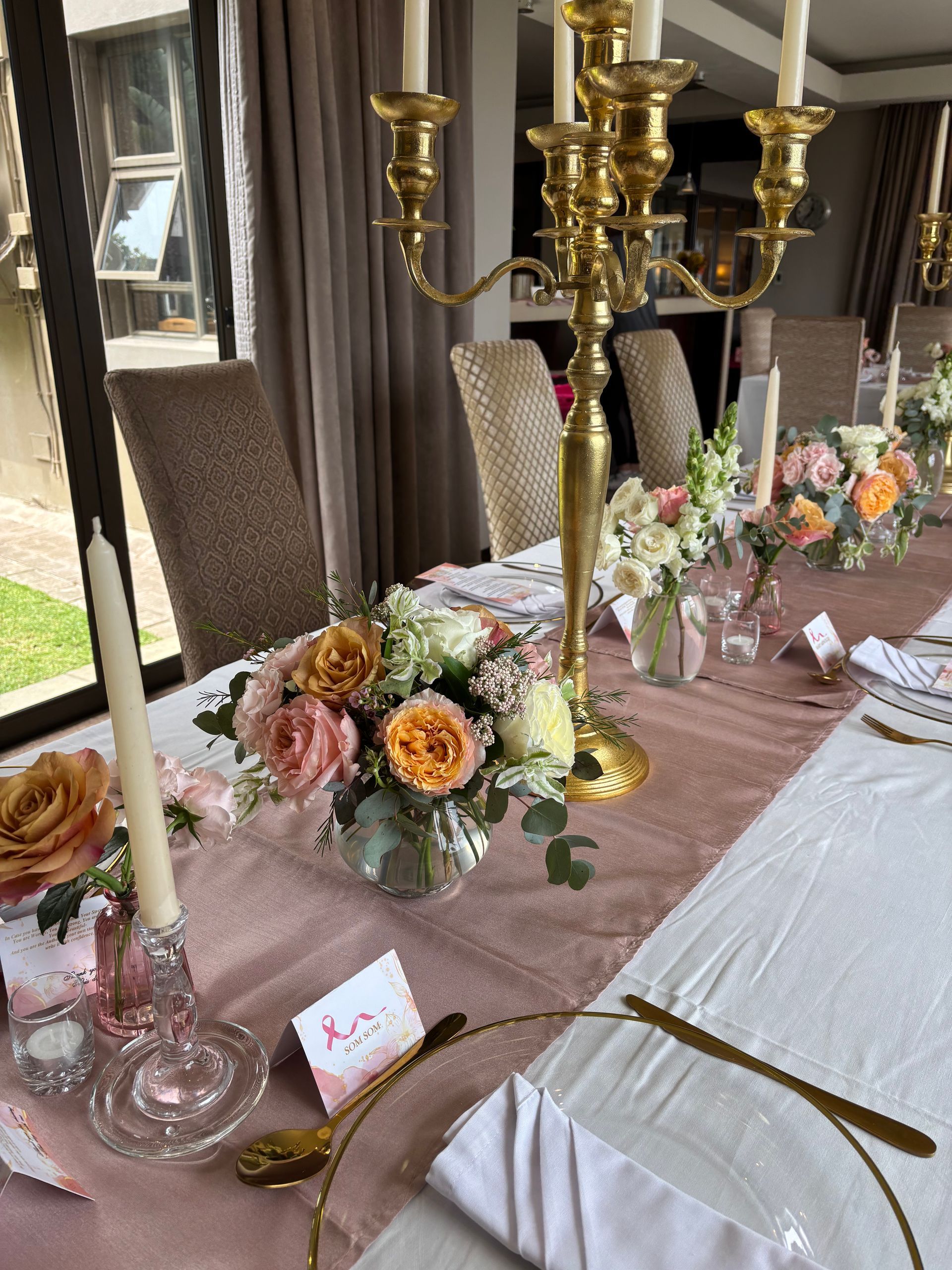 A formal dining table set for an event with dusty pink runners, floral arrangements, gold candelabras, and place settings.