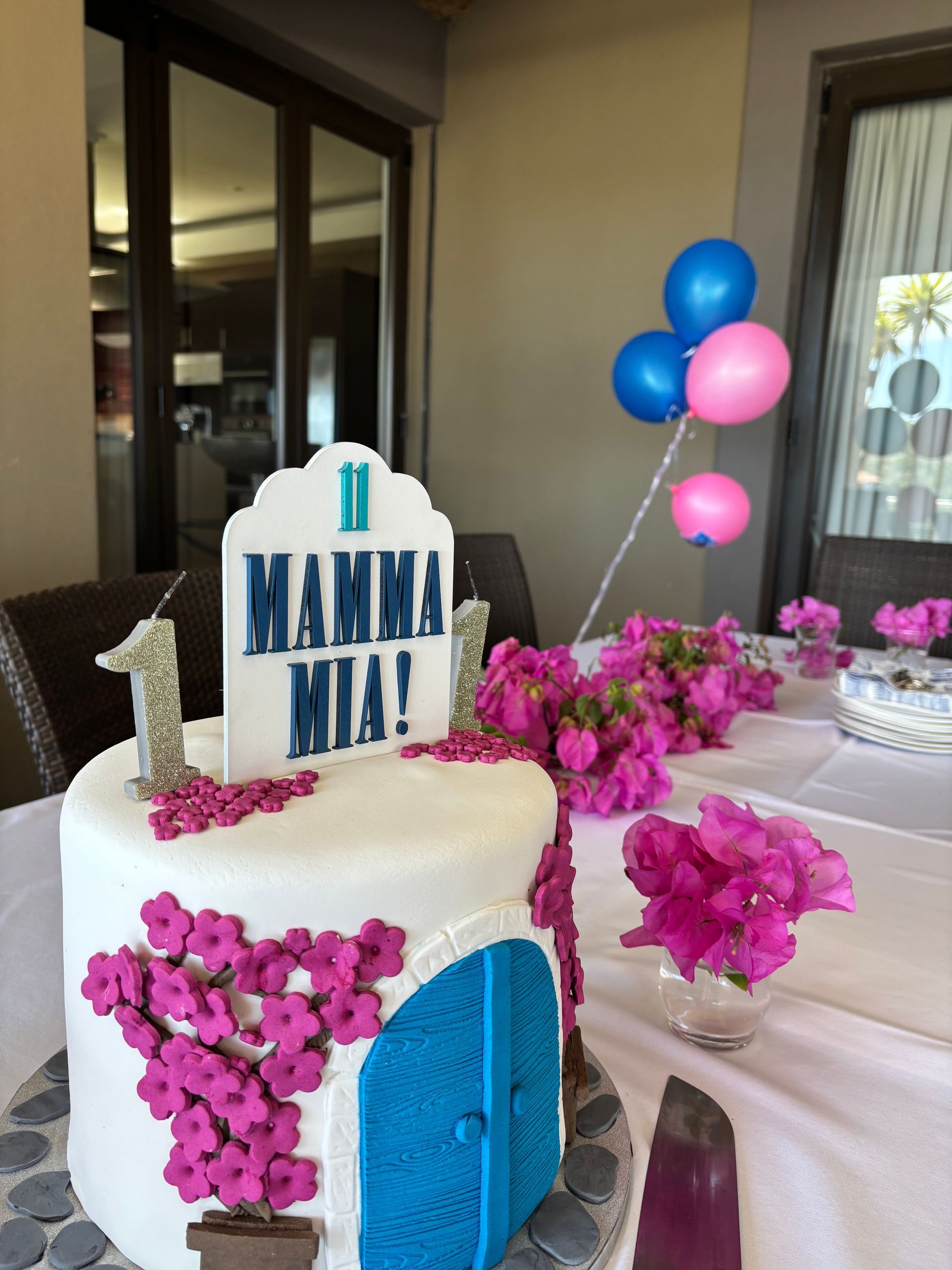 A Mamma Mia-themed birthday cake with a blue door, pink flowers, and a number one candle, set on a decorated table.