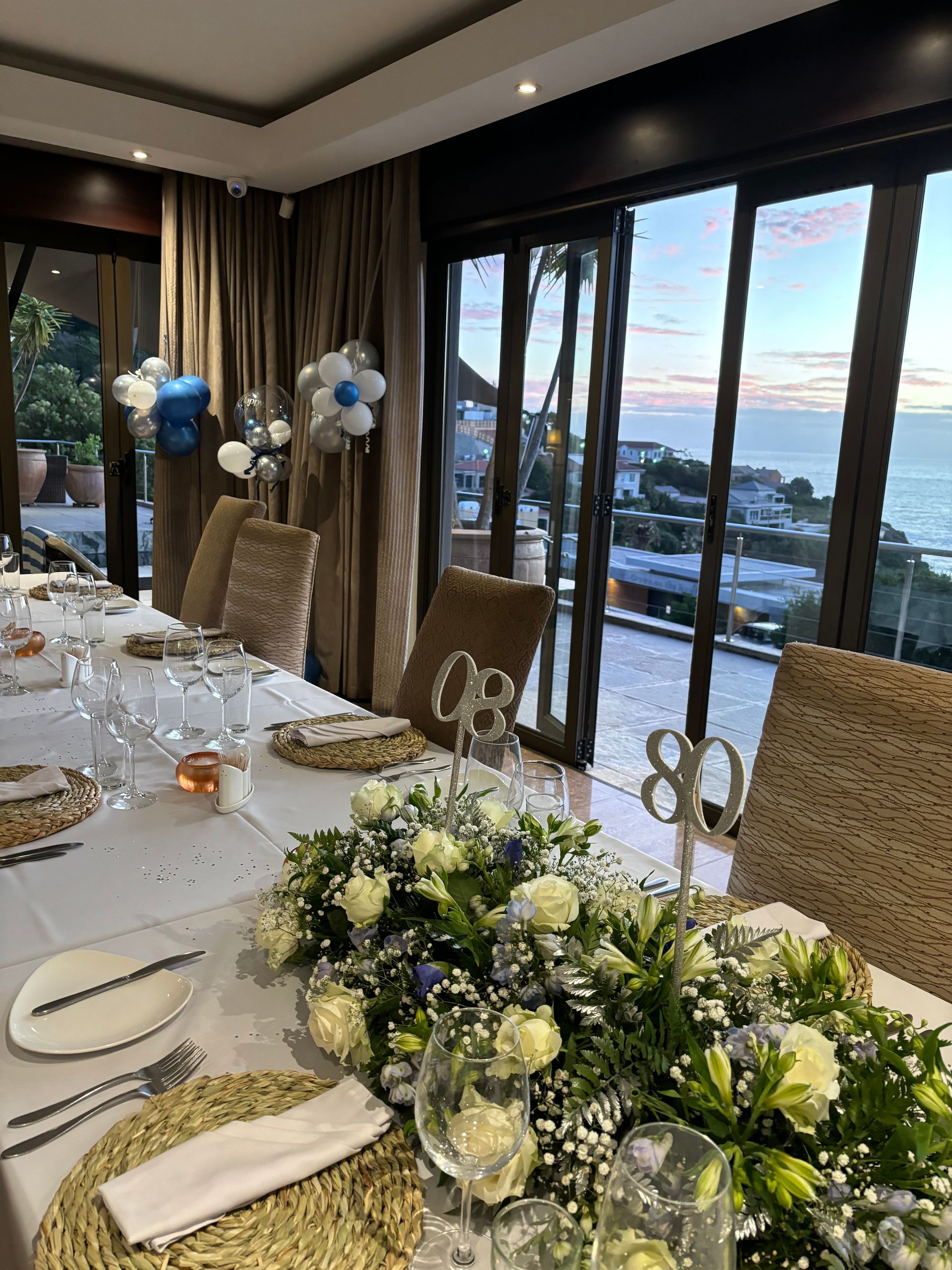 A long dining table set for an 80th birthday party with white floral centerpieces, in a room overlooking the ocean.