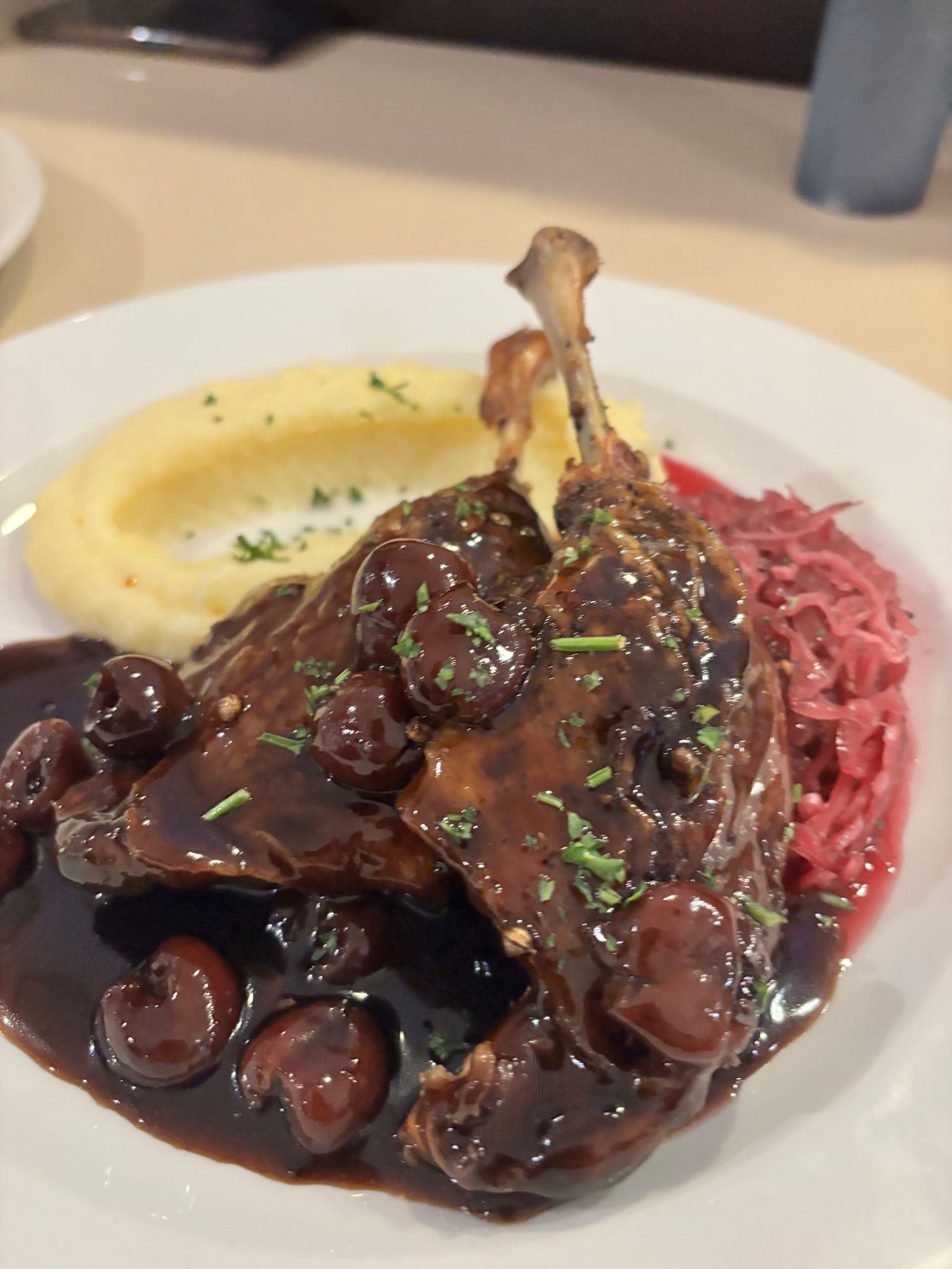 A plate of roasted duck legs topped with a dark cherry sauce, served with mashed potatoes and red cabbage.