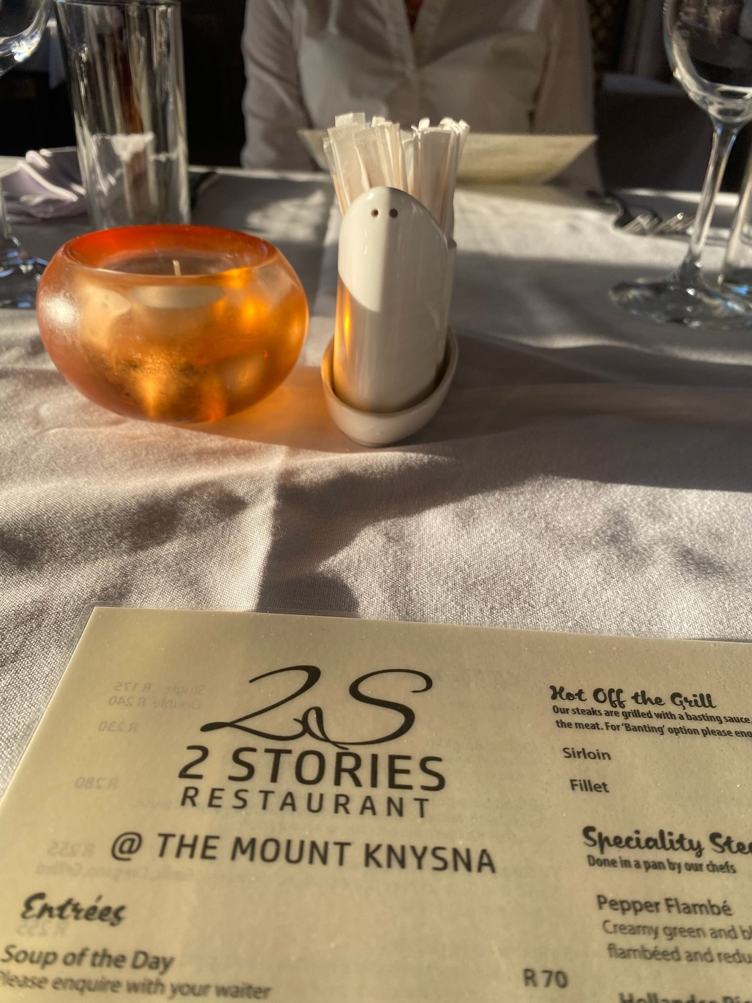 A menu for 2 Stories Restaurant at The Mount Knysna sits on a table with a lit orange candle and cutlery holder.