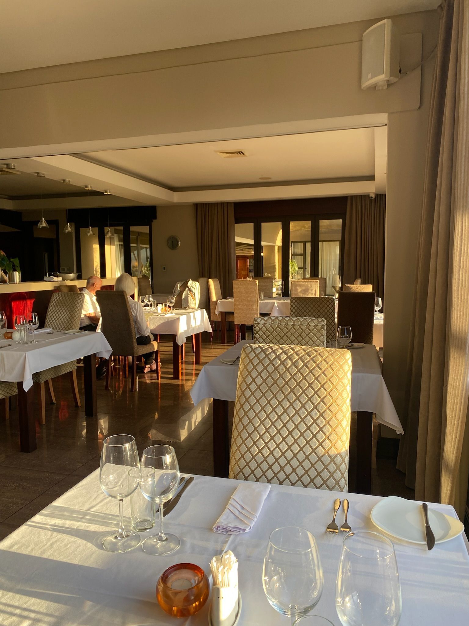 Sun-drenched dining room with white-clothed tables, chairs, and glasses, bathed in warm golden light.