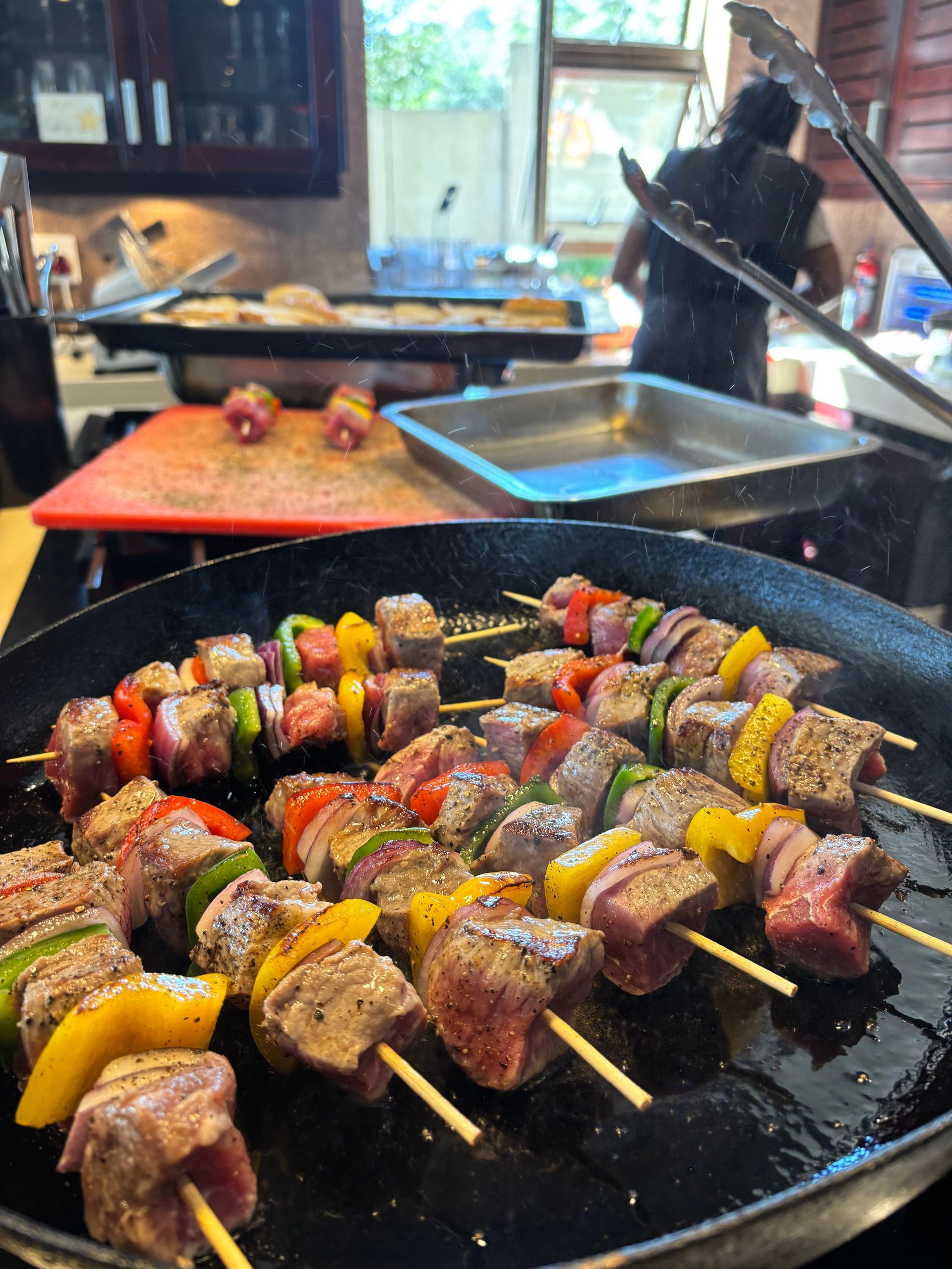 Skewers of meat and colorful bell peppers cooking in a frying pan on a kitchen stove, with a person preparing food nearby.