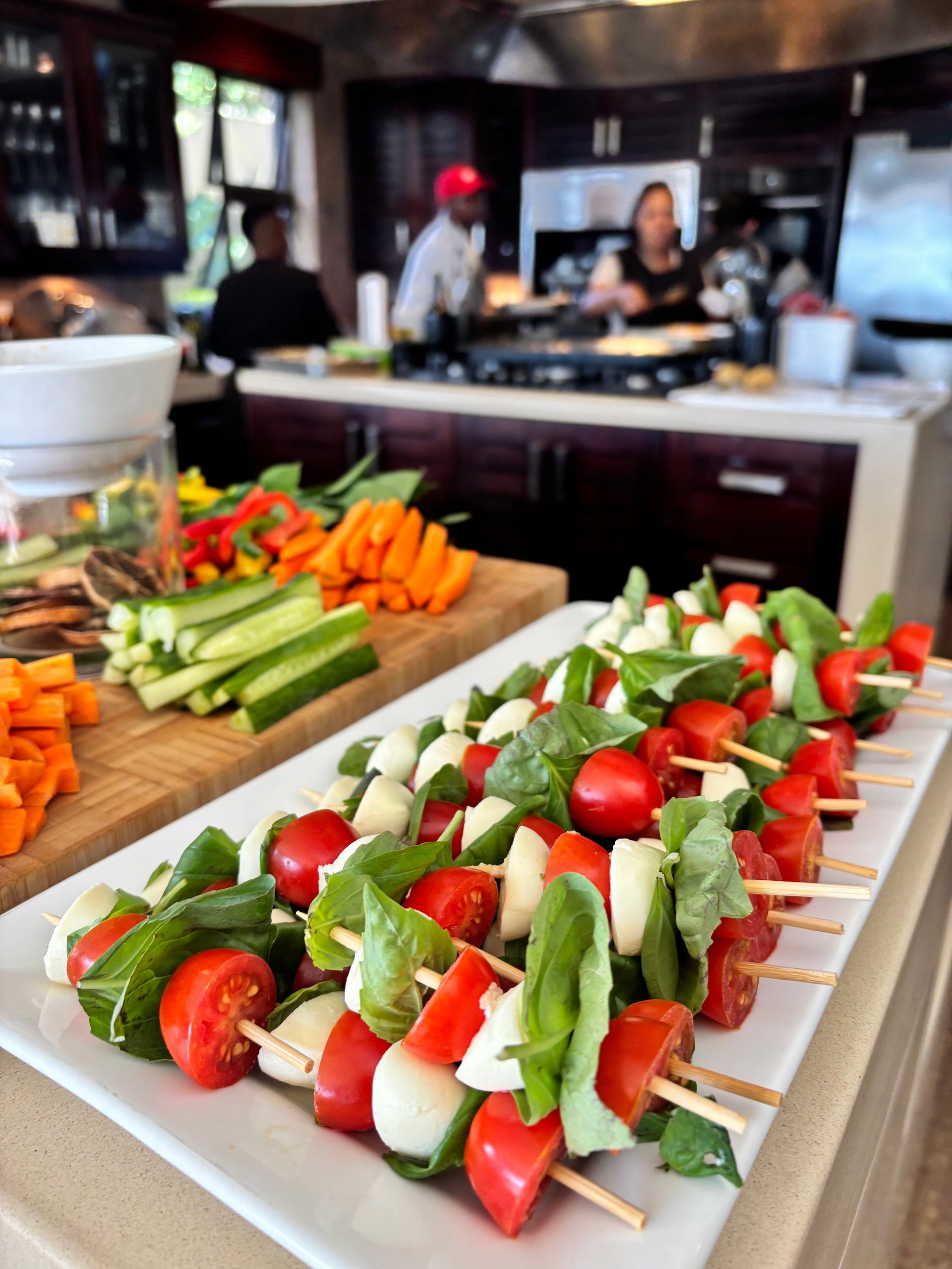 A tray of tomato, mozzarella, and basil skewers sits in the foreground of a kitchen, with staff preparing food behind it.