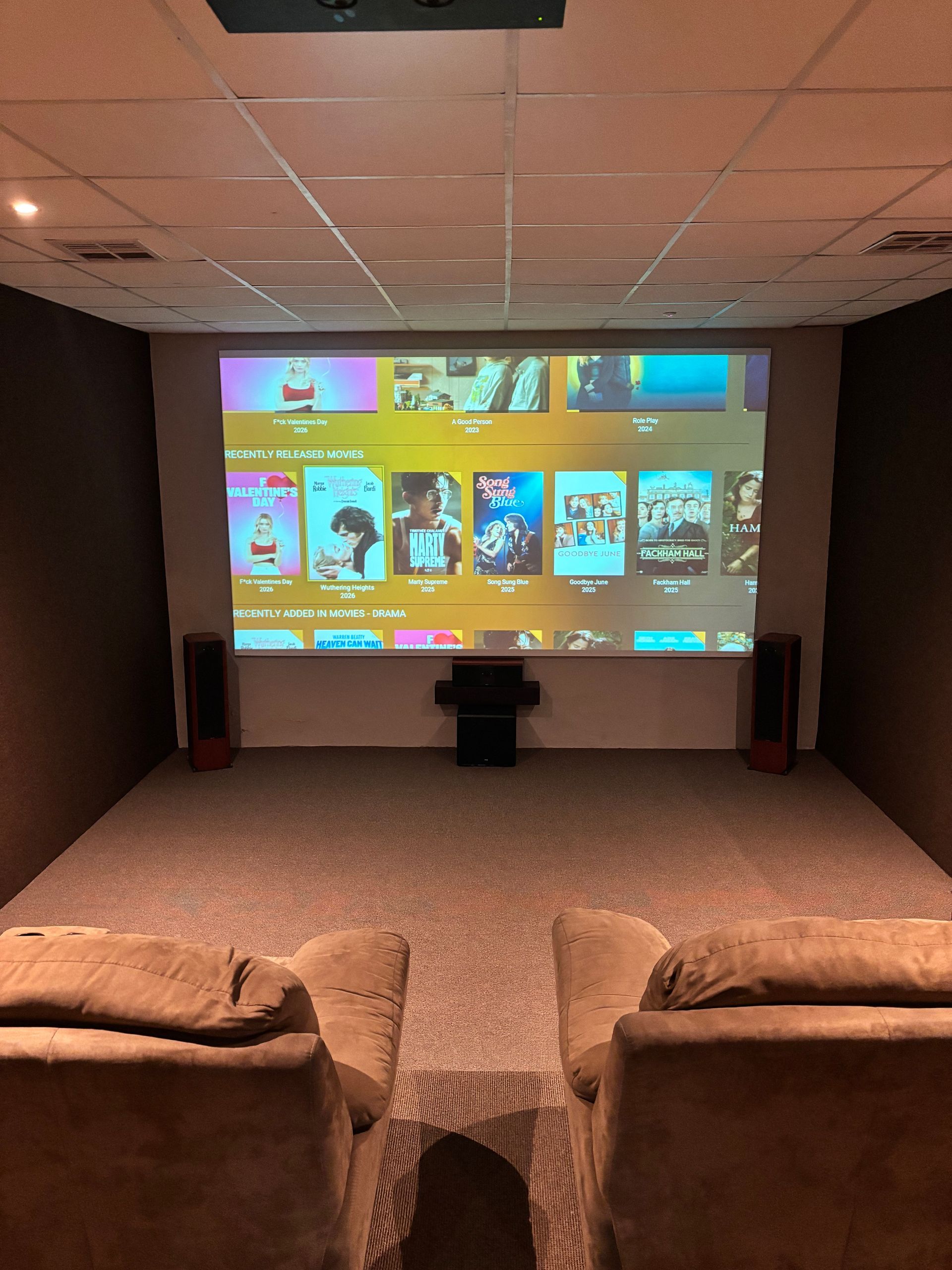 A cozy home theater with two brown recliners facing a large projection screen displaying a digital movie menu.
