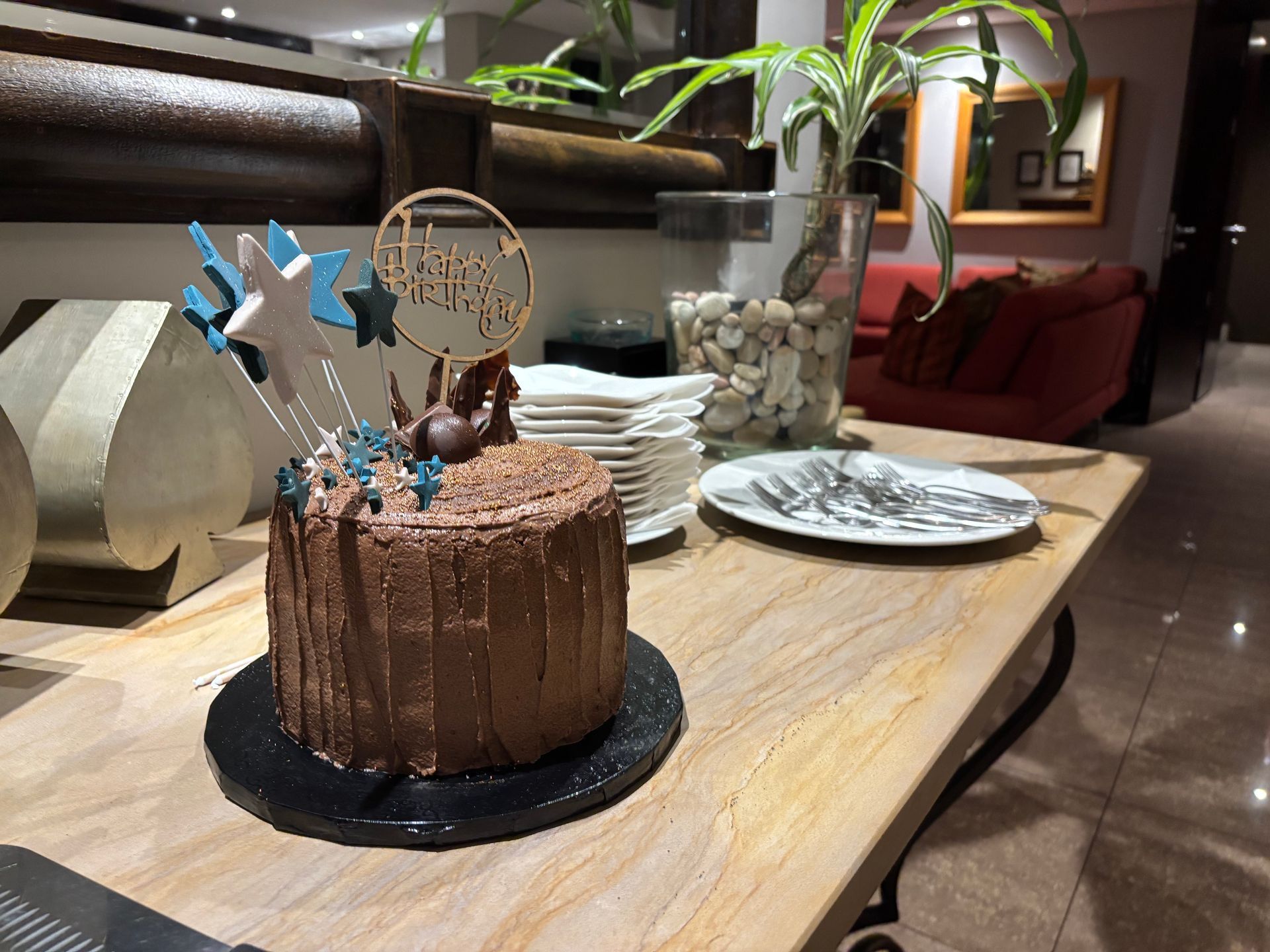 A chocolate drip cake with star toppers and a gold cake topper on a wooden table, next to plates and a plant.