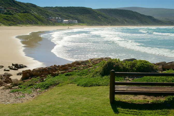 Beach scene with ocean waves, tan sand, green hills, and a wooden bench in the foreground.