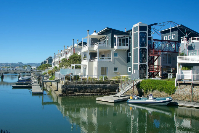 Waterfront houses with docks and a bridge in the background, under a blue sky.