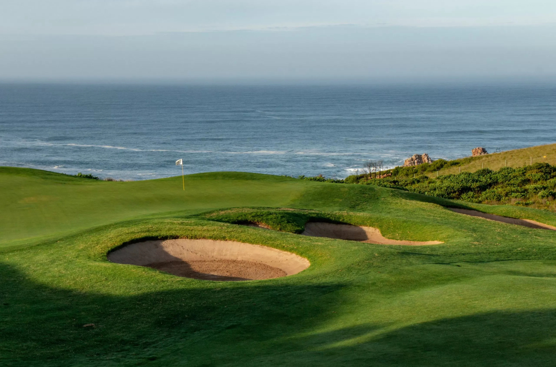 Golf course green with sand traps, ocean backdrop, and a flag.