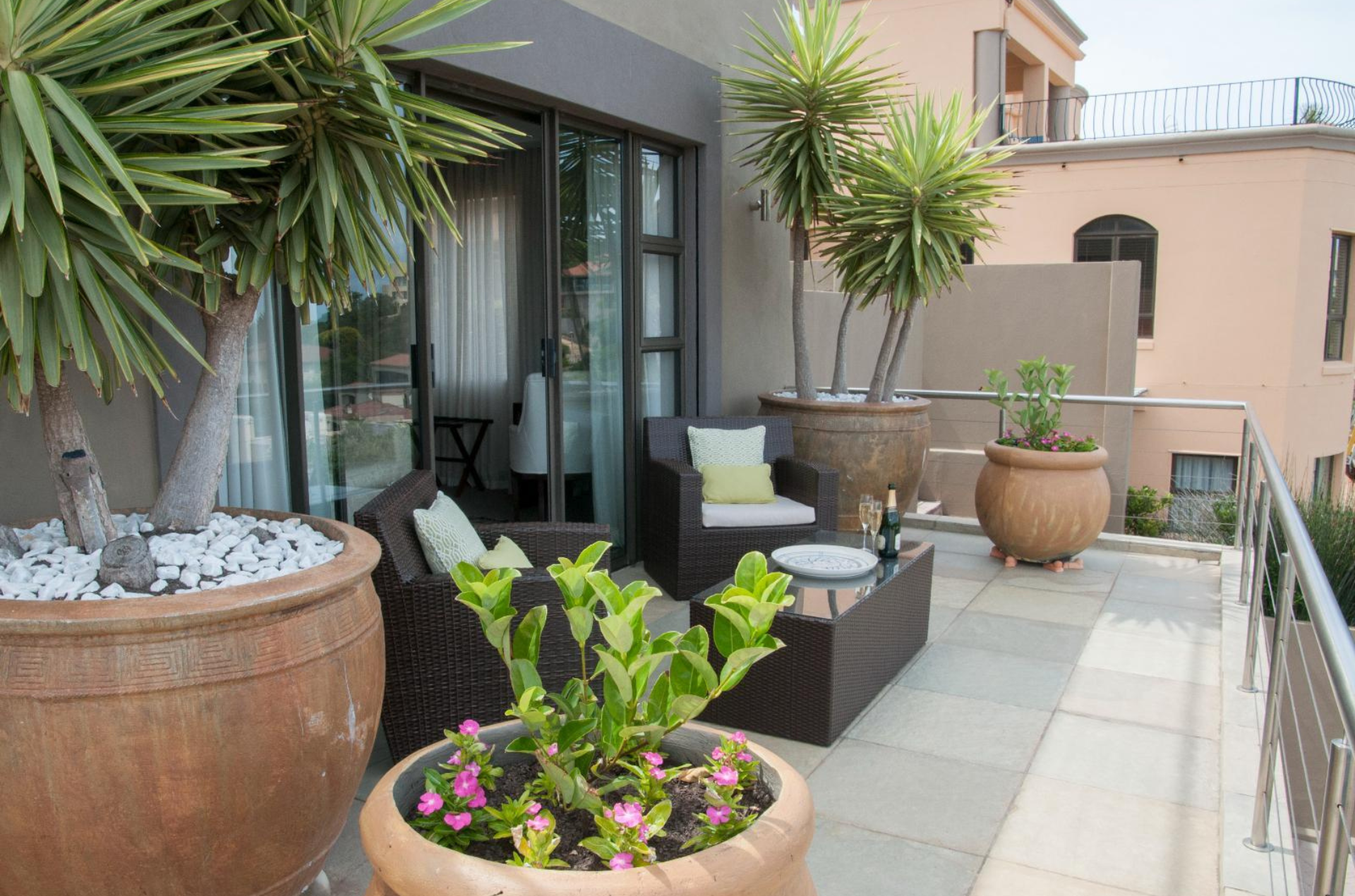 Balcony with potted plants and seating. Sliding glass door leads to a room. Beige tones.