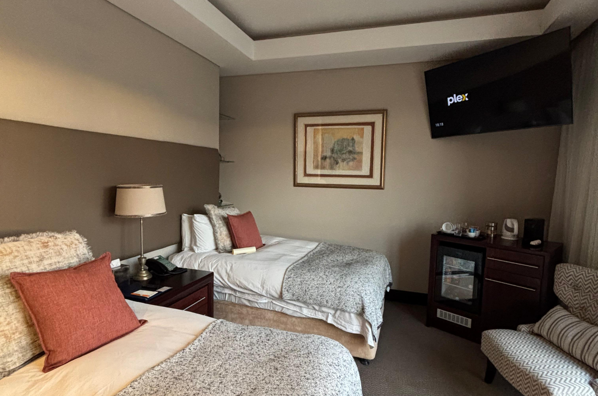 Two twin beds in a hotel room with TV, nightstands, chair, and artwork. Muted color palette.