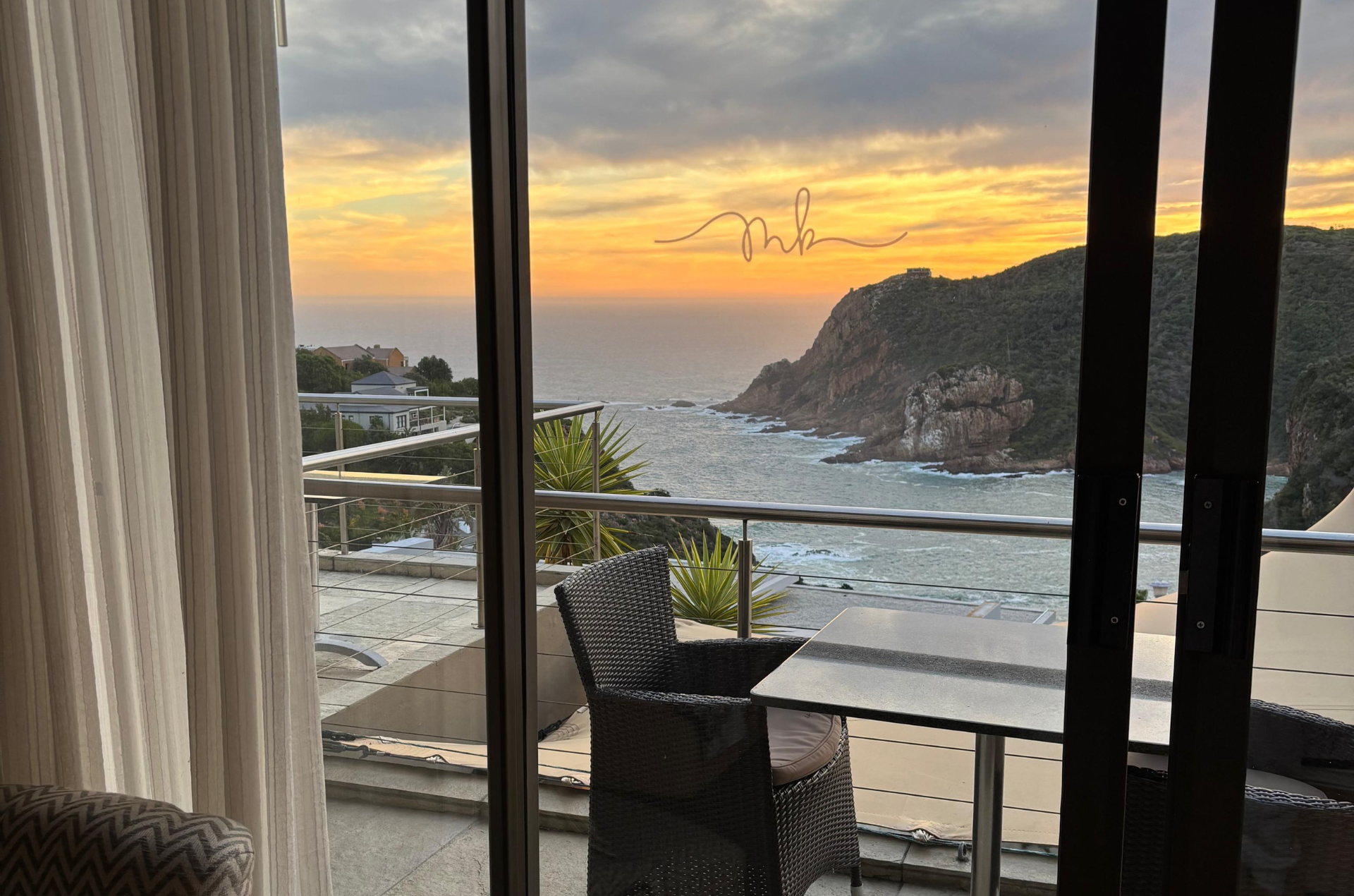 Sunset view through a window, overlooking a bay, with a table and chair on a balcony.