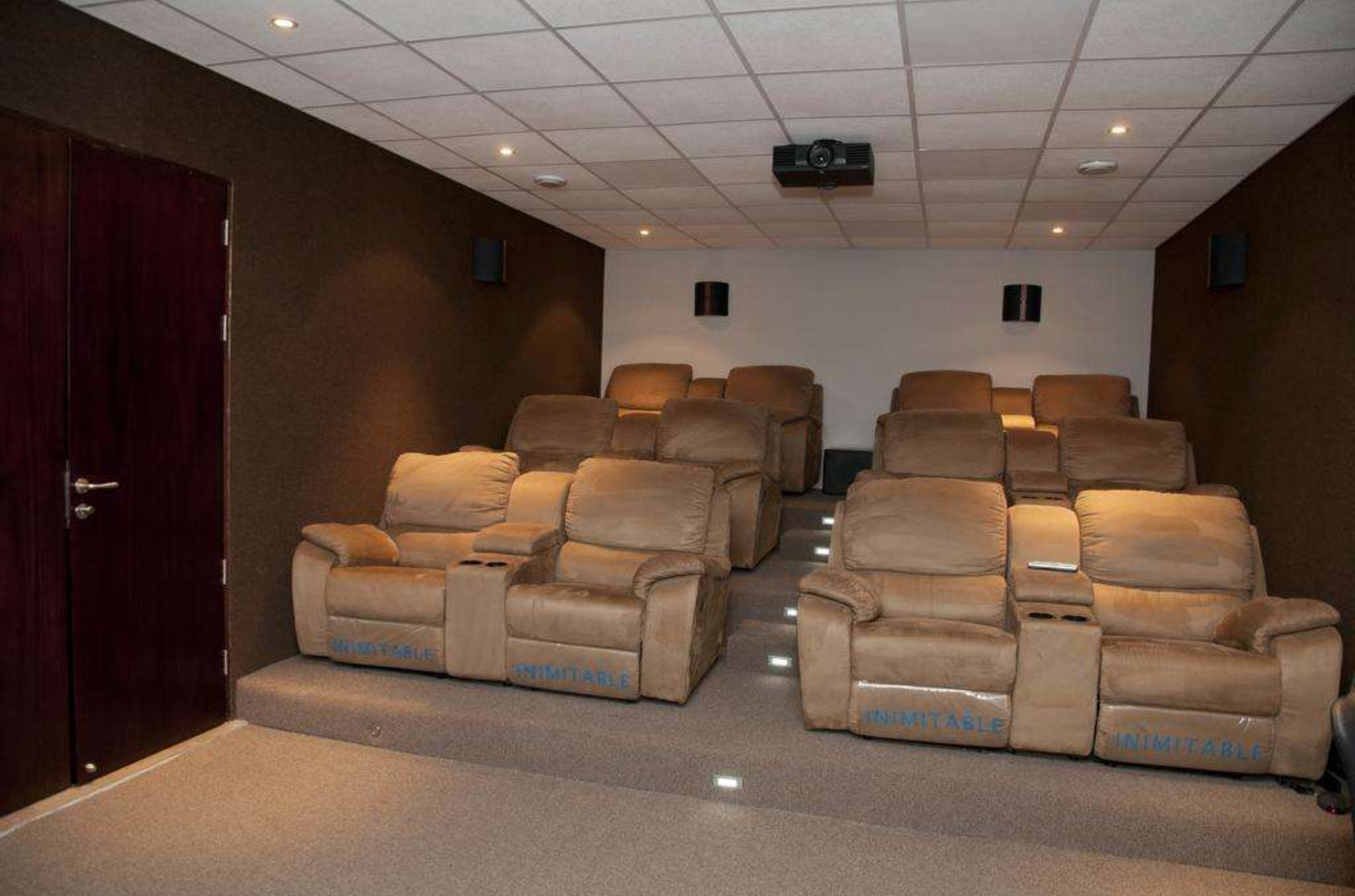 A small home theater with two rows of beige reclining chairs on tiered carpeted platforms and a projector on the ceiling.