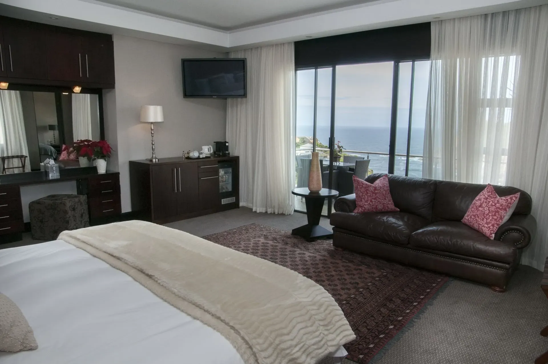 Hotel room with a bed, seating area, and ocean view. Dark furniture, neutral walls, and a patterned rug.