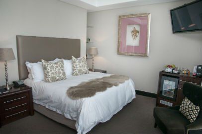 Bedroom with a bed, nightstands, chair, mini-fridge, wall art, and TV. Neutral colors.