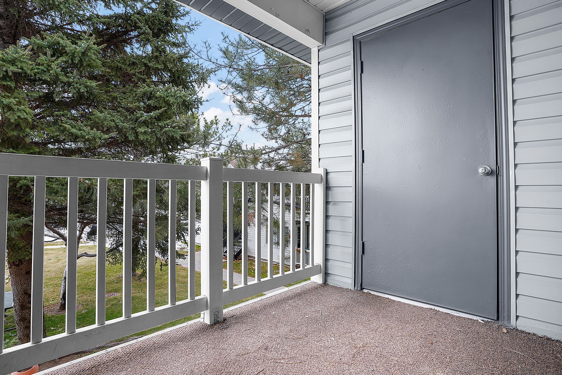 Balcony with gray door, railing, and view of trees and sky.