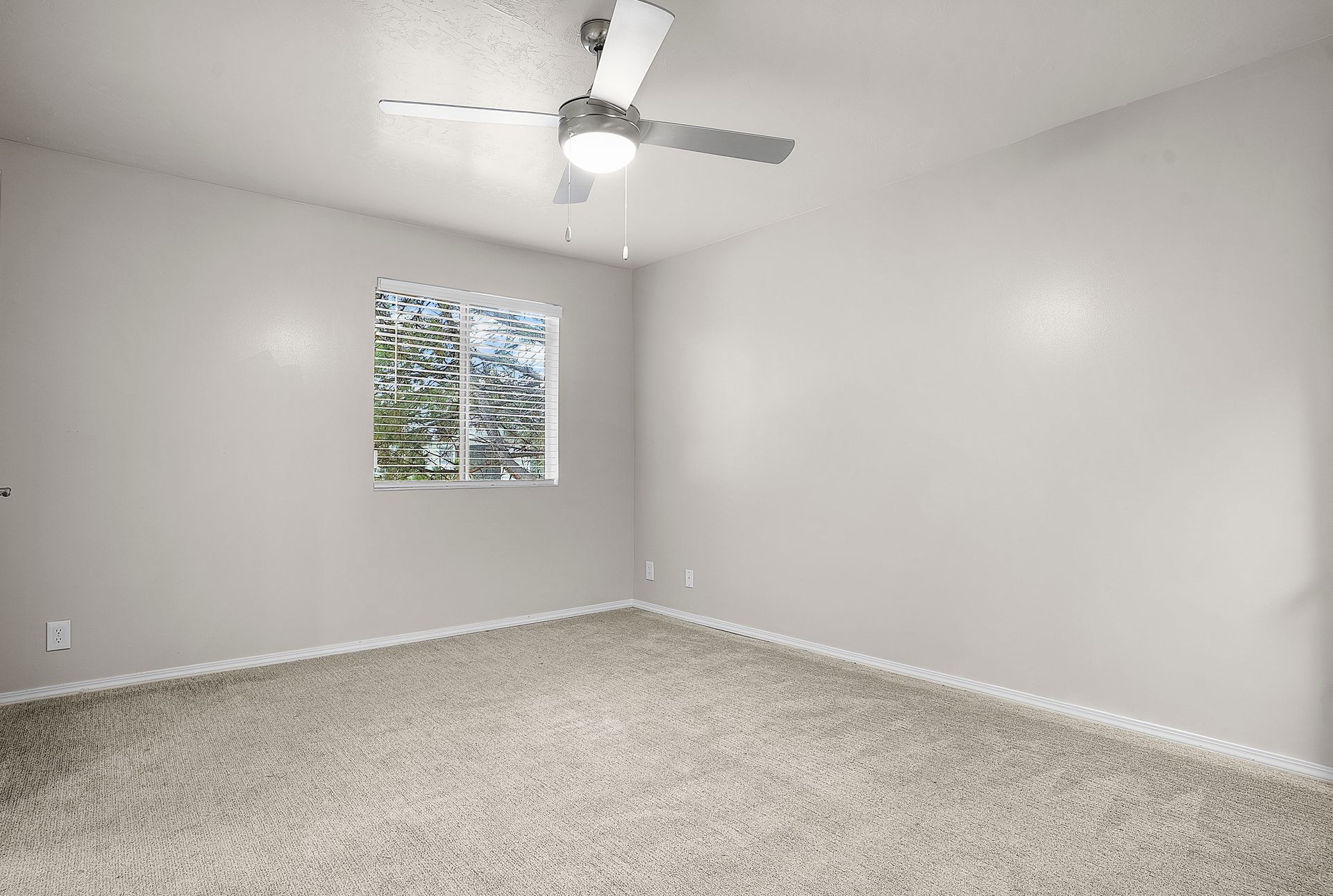 Empty bedroom with light gray walls, window, ceiling fan, and beige carpet.