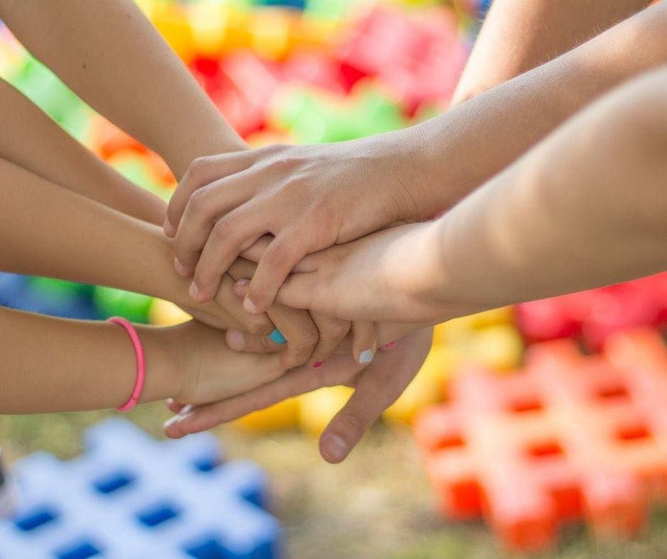 Un gruppo di mani sovrapposte a formare un cerchio, con sullo sfondo blocchi di costruzione geometrici e colorati sfocati.