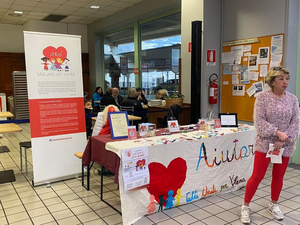 In una stanza luminosa, una donna è in piedi accanto a un tavolo informativo di beneficenza con uno striscione a forma di cuore con la scritta "Aiutar".