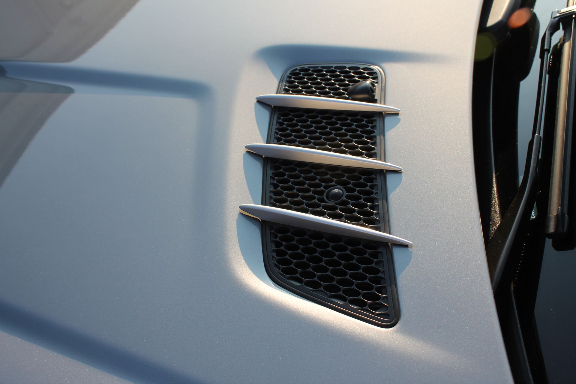 Black car hood vent with three horizontal silver bars and honeycomb mesh.