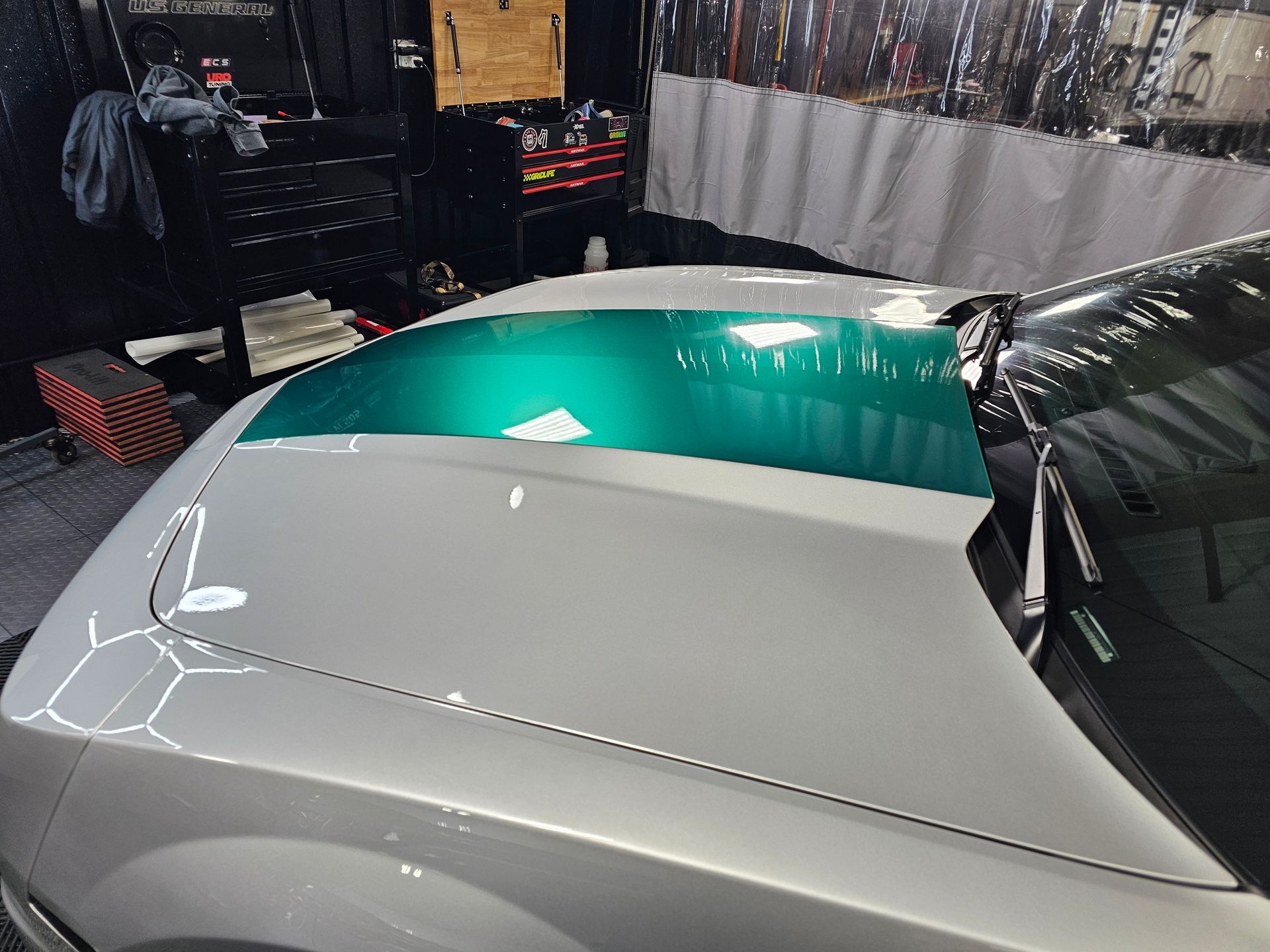 Car hood partially covered in silver and teal vinyl wrap, in a shop.