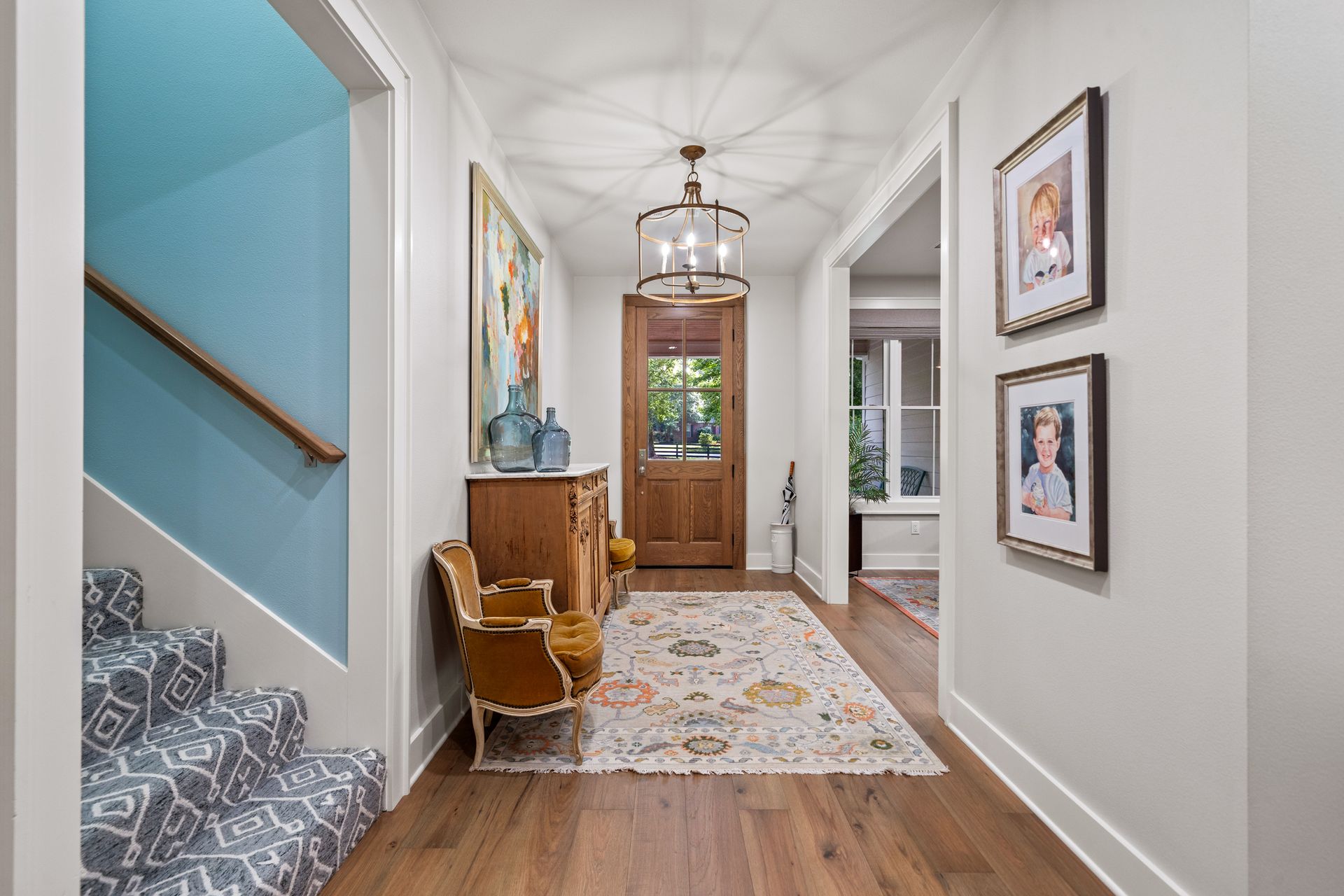 An elegant entryway features hardwood floors, a patterned rug, a wooden door, a staircase, and wall-mounted art.