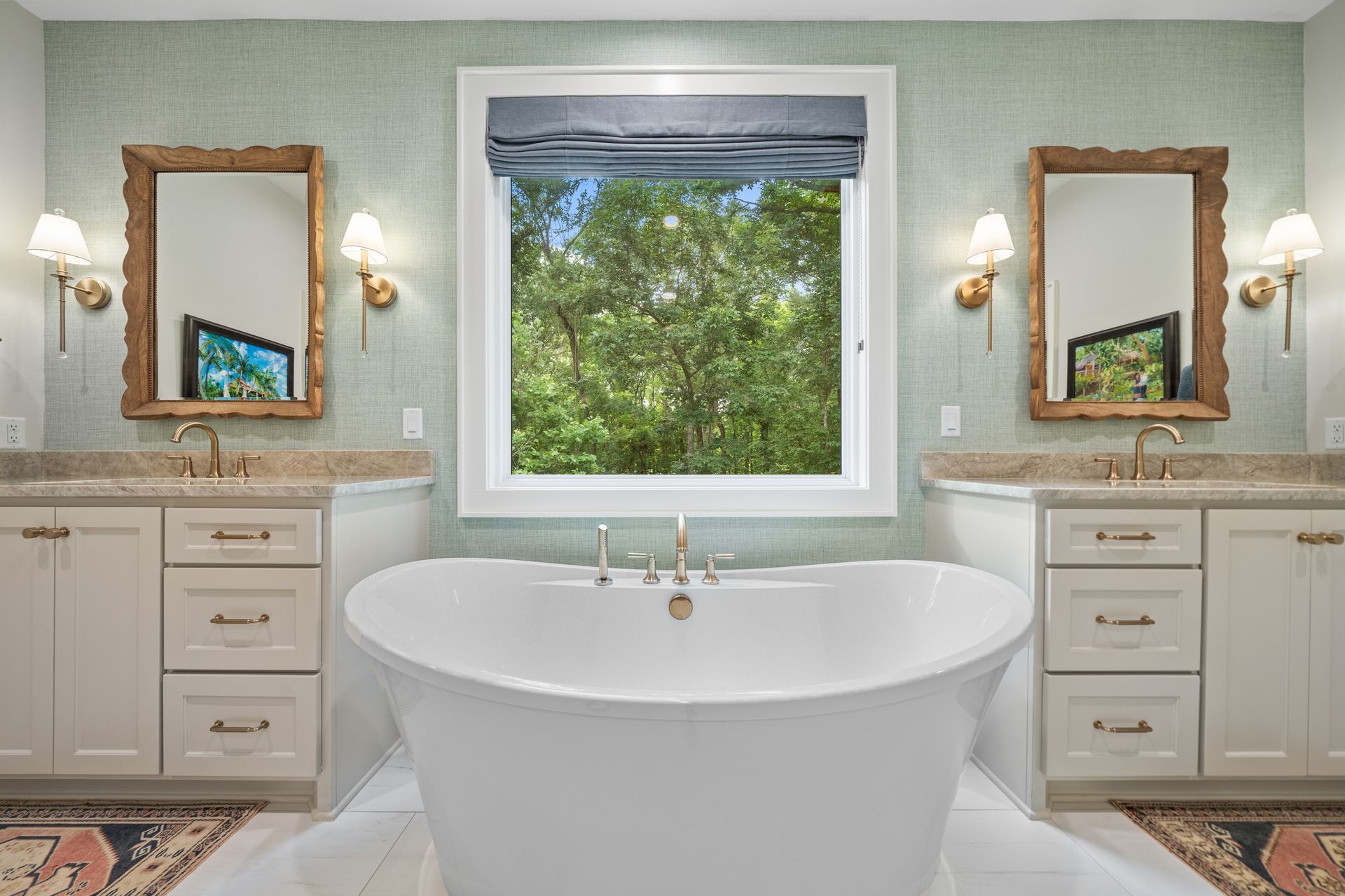 A bathroom with a freestanding white tub centered before a window, flanked by two vanities with mirrors and gold sconces.