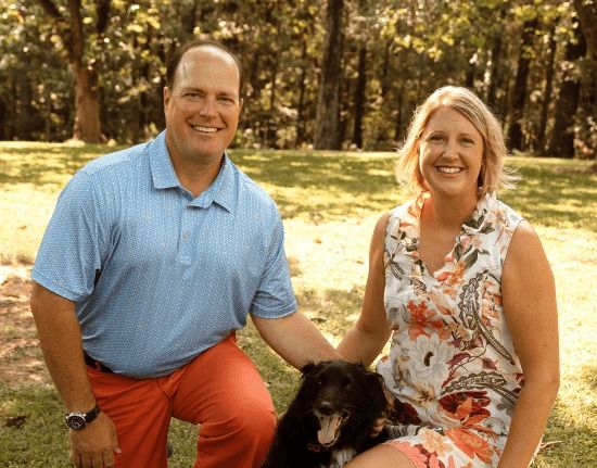 A couple smiling while kneeling in a grassy field with a small black dog between them.