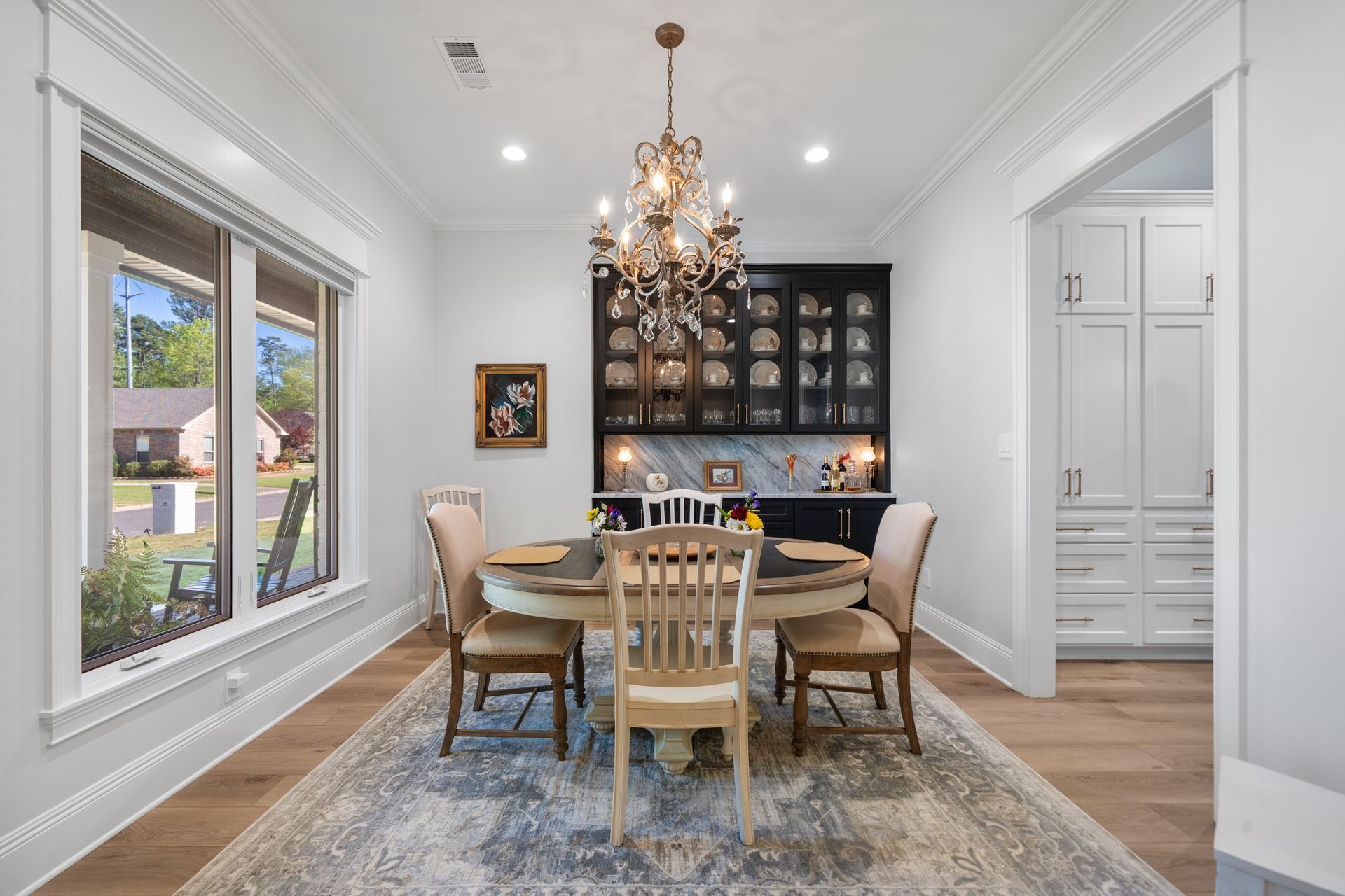Dining room with a round table, four chairs, a crystal chandelier, and a black built-in hutch on a patterned rug.