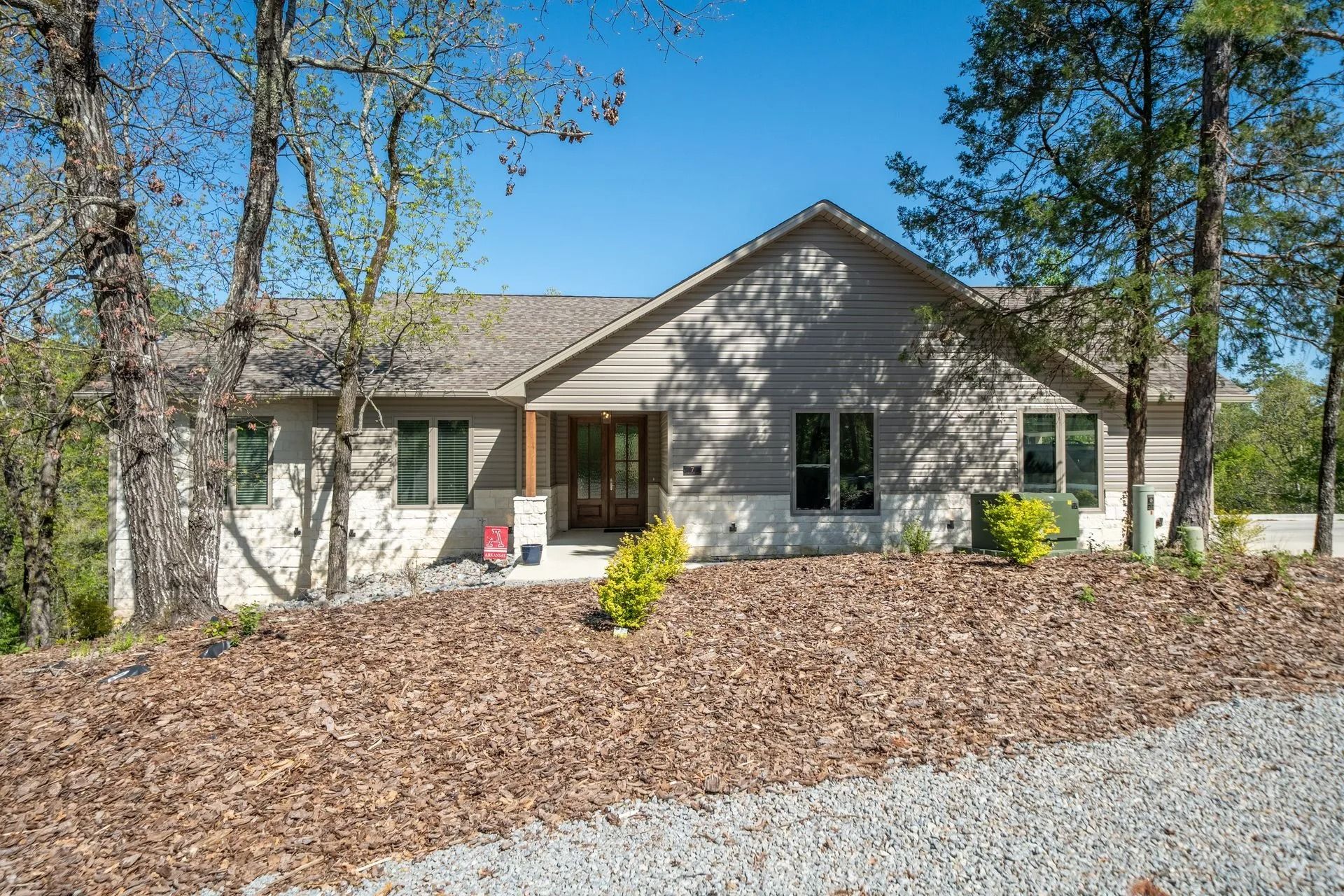 A single-story suburban house with light gray horizontal siding, a stone base, and a gravel driveway in a wooded area.