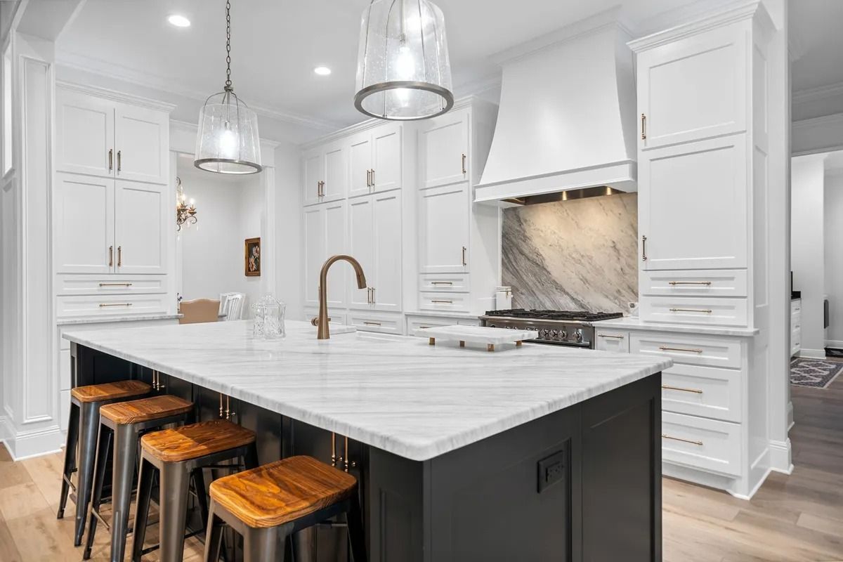 A modern kitchen featuring a dark island with marble countertop, white cabinetry, a marble backsplash, and hanging lights.