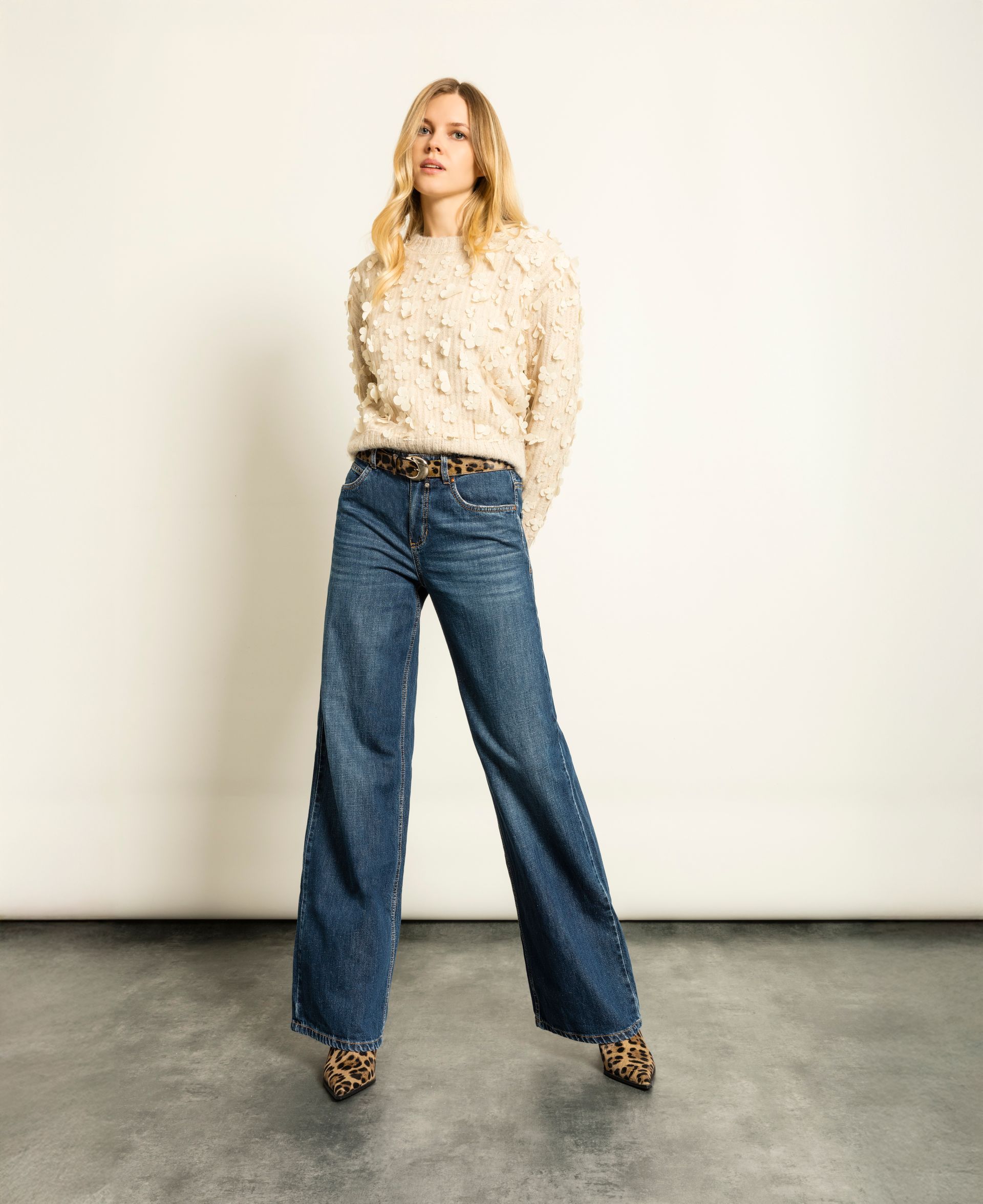 Eine Frau in einem strukturierten Pullover, einer weit geschnittenen Jeans, einem gemusterten Gürtel und spitzen High Heels posiert im Studio.
