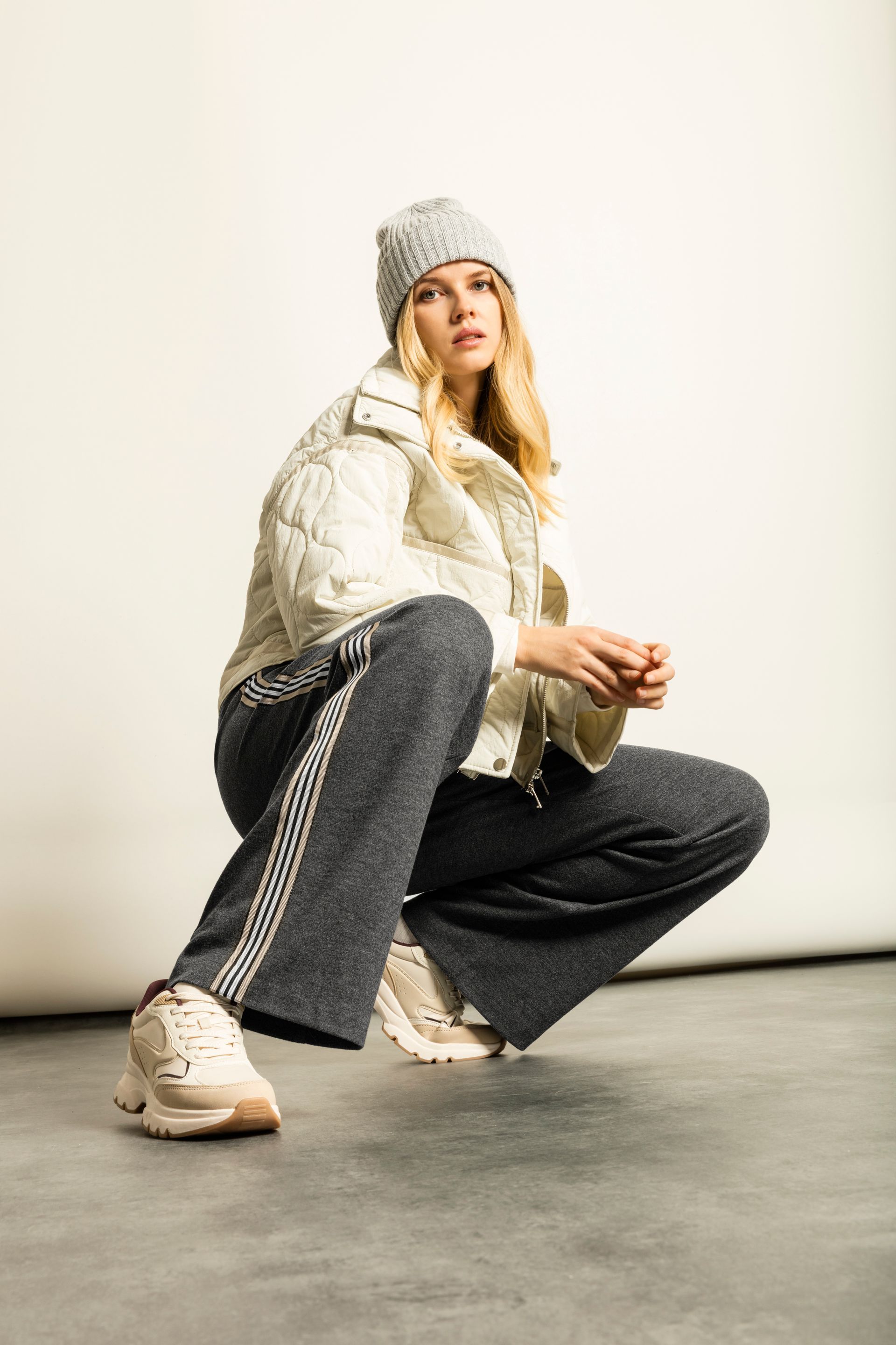 Frau in Steppjacke, grauer Jogginghose, Mütze und Turnschuhen macht Kniebeugen.