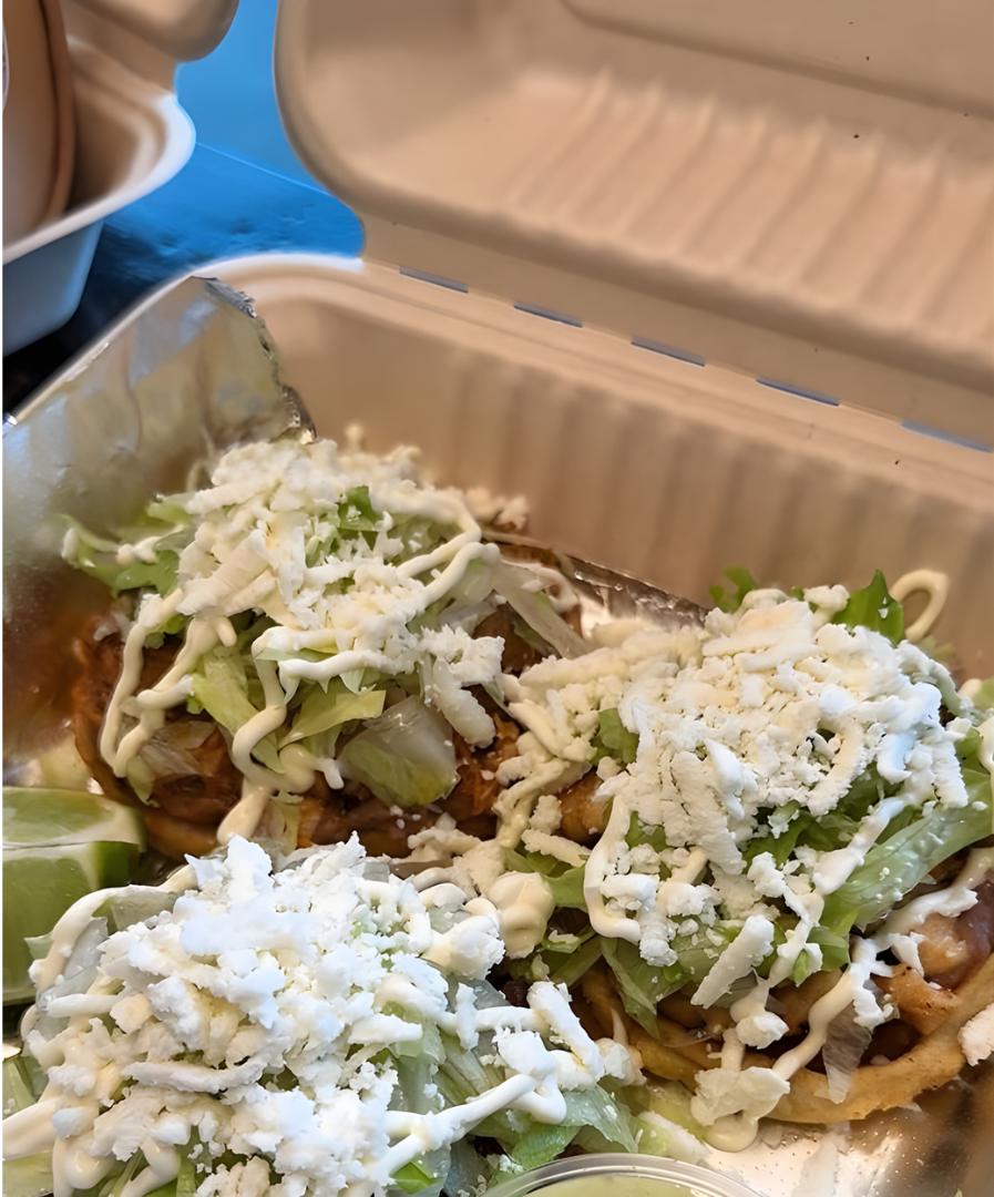 Three tostadas topped with shredded lettuce, crumbled cheese, and a drizzle of crema inside a takeout container.