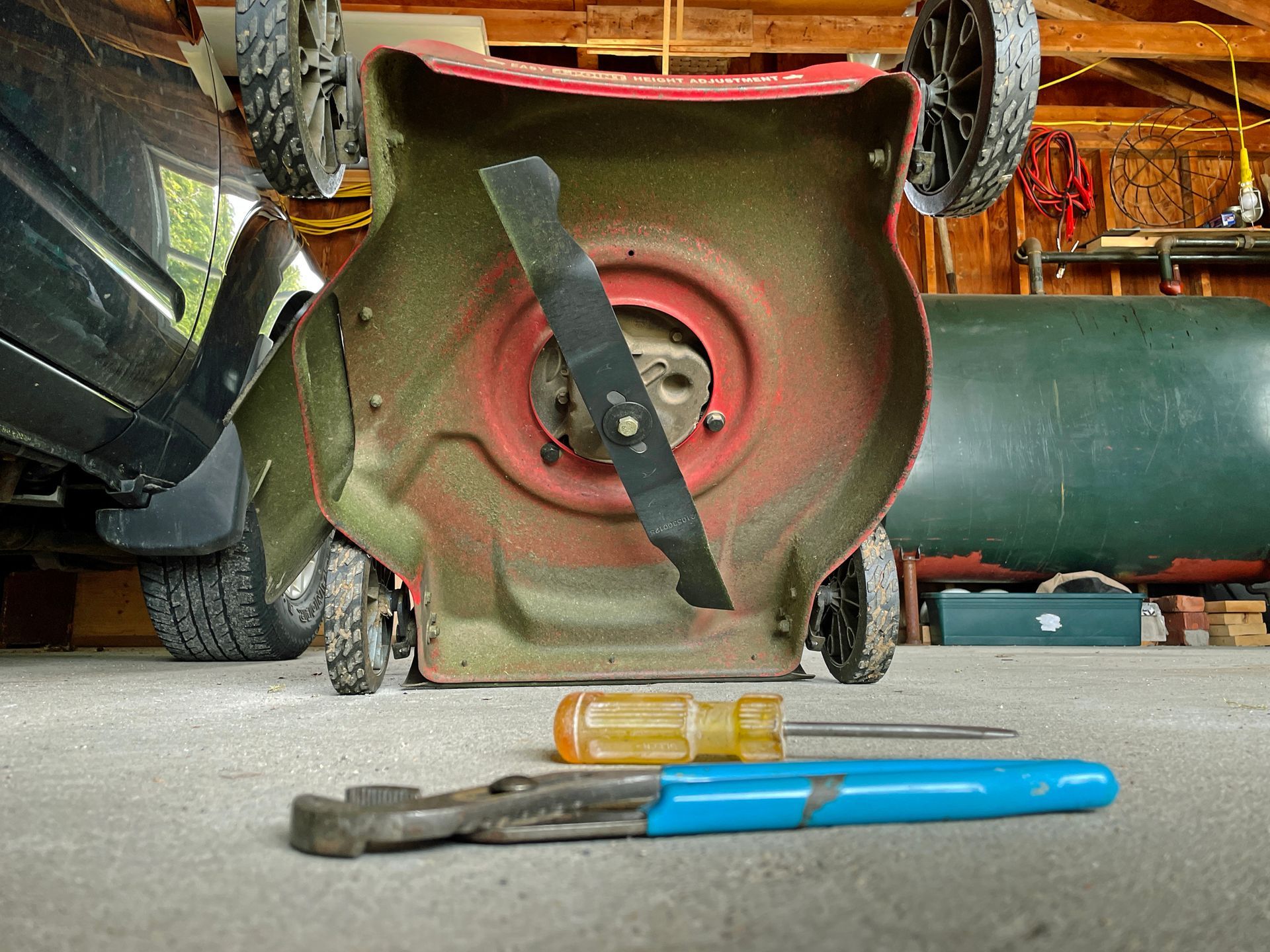 Lawnmower upside down, blade exposed, with wrench and screwdriver on ground in a garage.