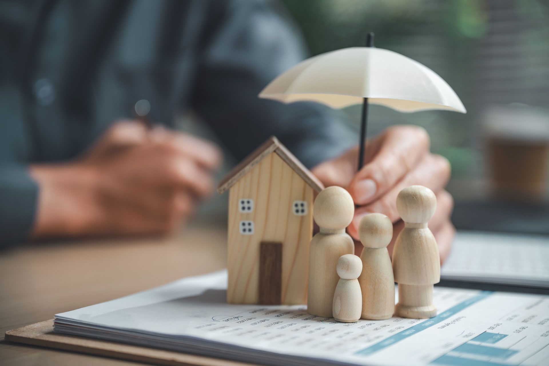 Person holding a small umbrella over a wooden house and family figurines, symbolizing insurance protection.