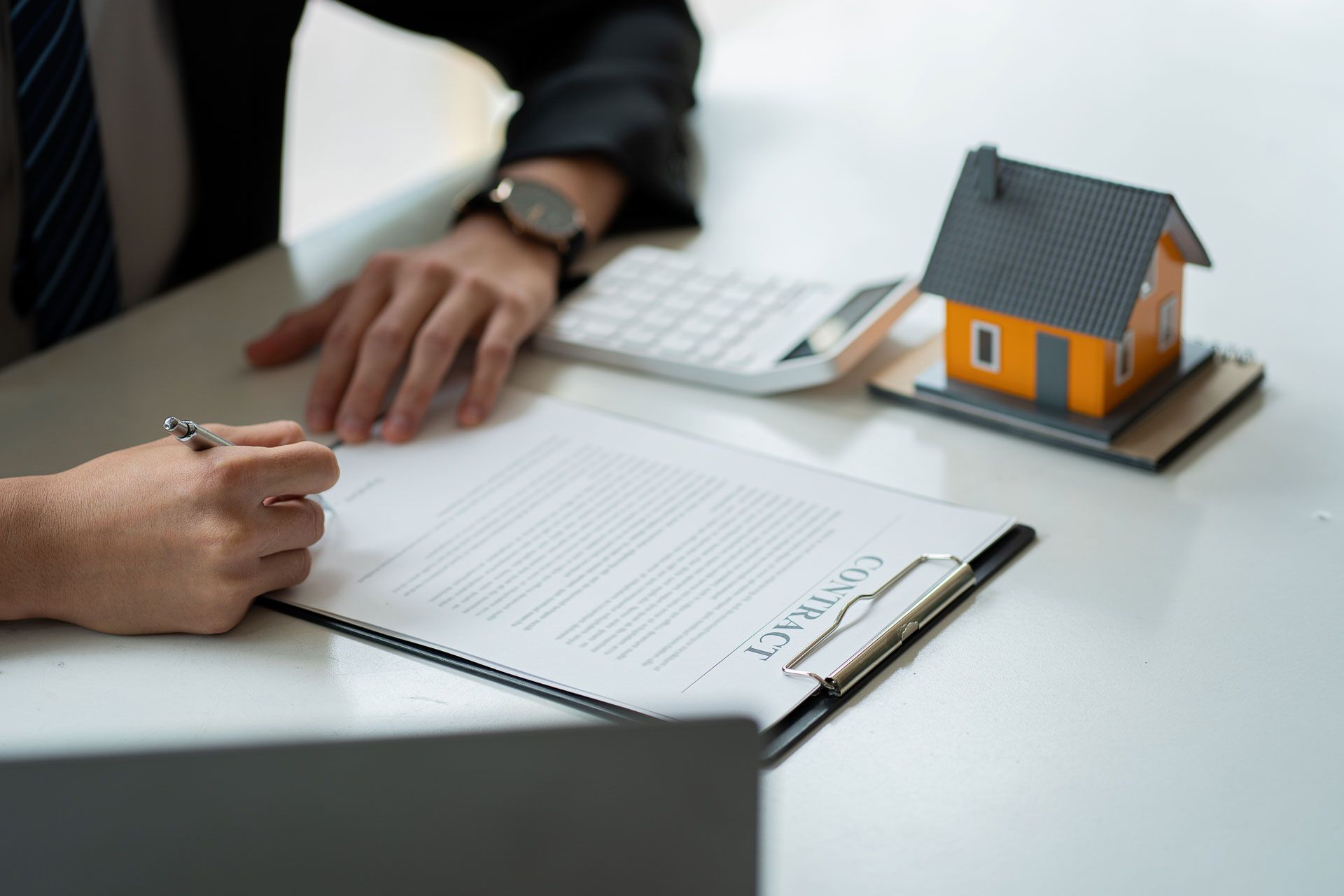 Person signing a contract, model house and calculator on a desk.