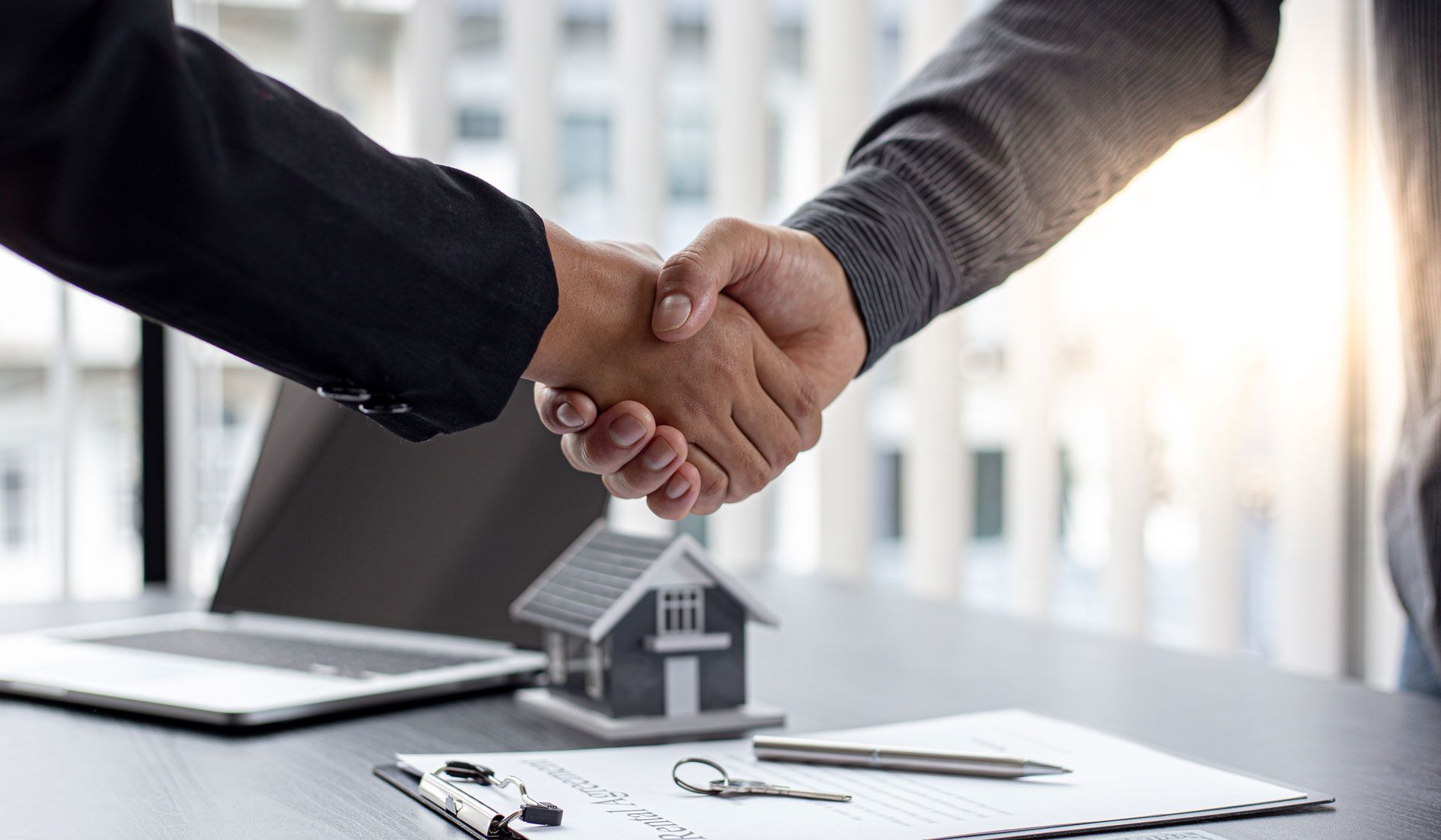 Two people shaking hands over a miniature house, documents, and a laptop.