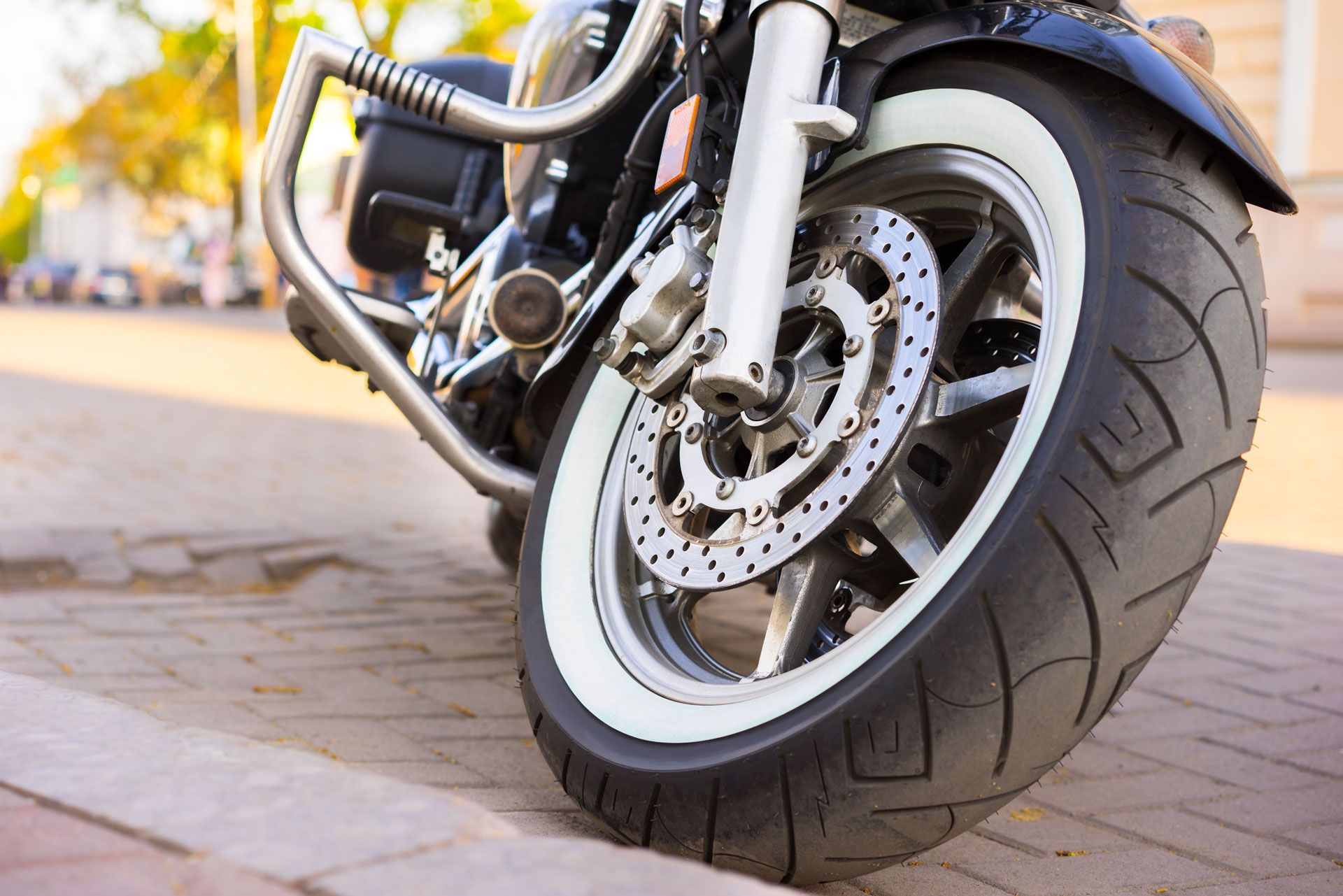 Close-up of a black motorcycle front wheel with a white sidewall tire, parked on a brick-paved road.