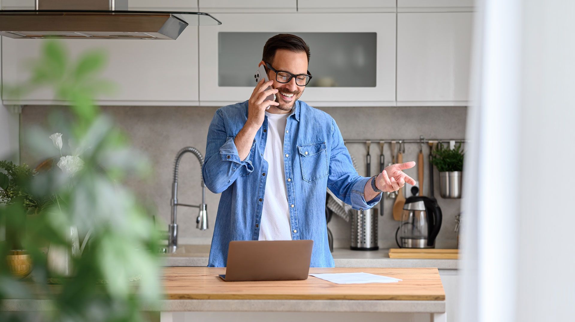 Man on phone in kitchen, smiling, gesturing, and working on laptop.