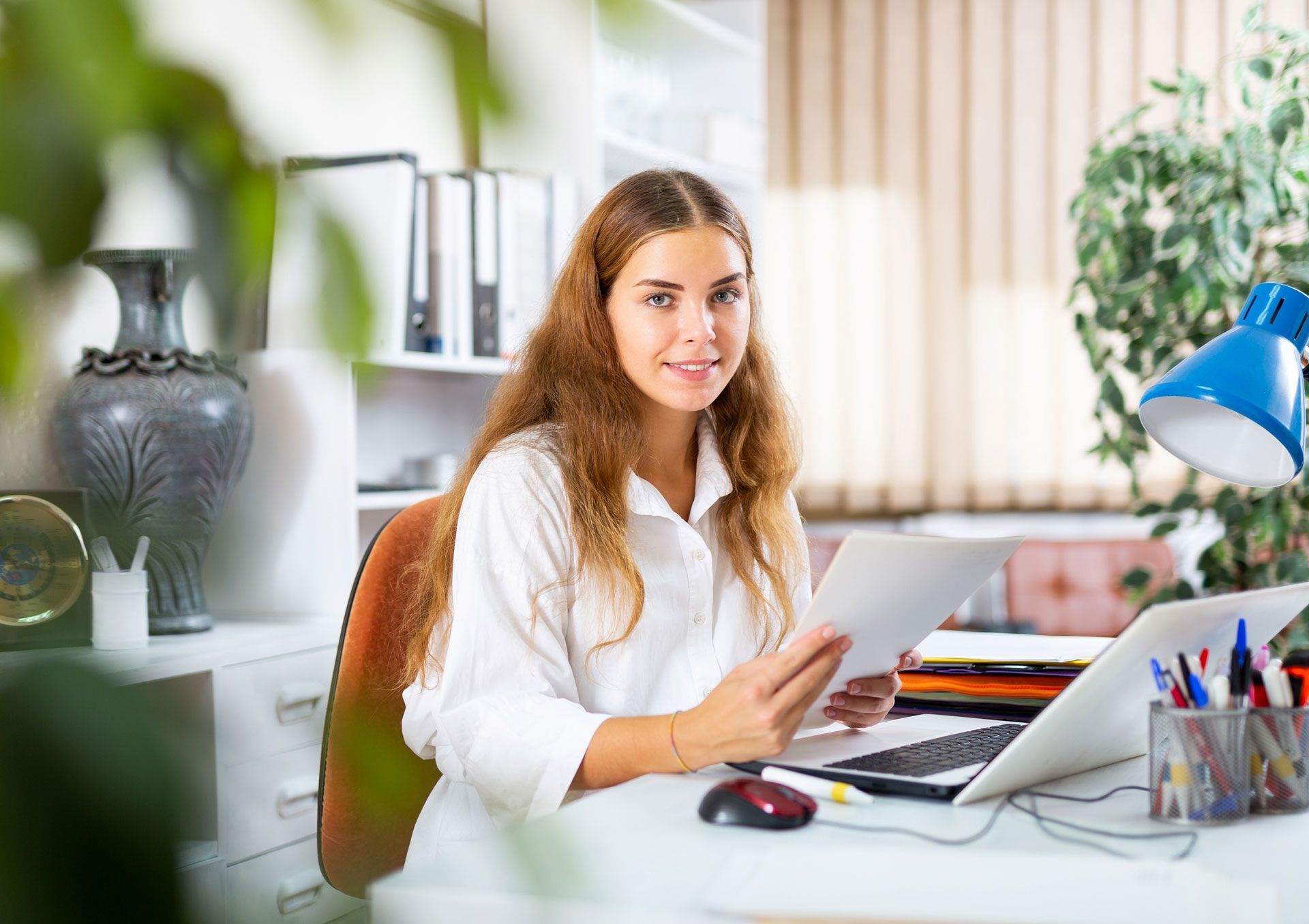 Woman in white shirt, seated at a desk, holding papers, smiling, with a laptop, office setting.