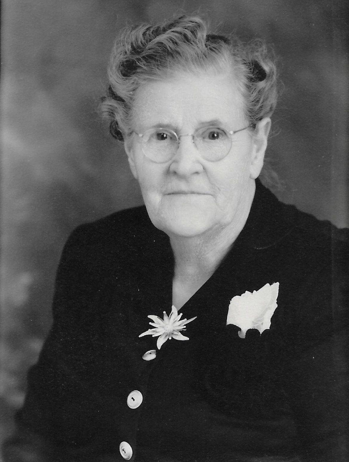 Portrait of an elderly woman in a dark suit with a flower brooch, wearing glasses and a composed expression