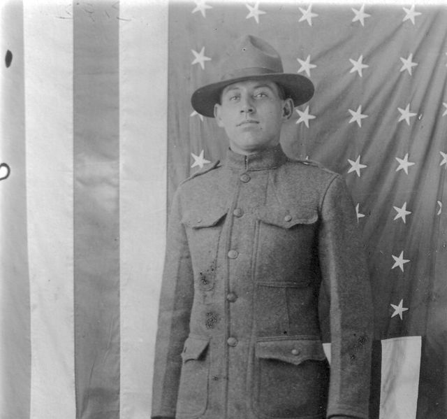 Soldier in uniform standing before American flags, wearing a wide-brimmed hat.