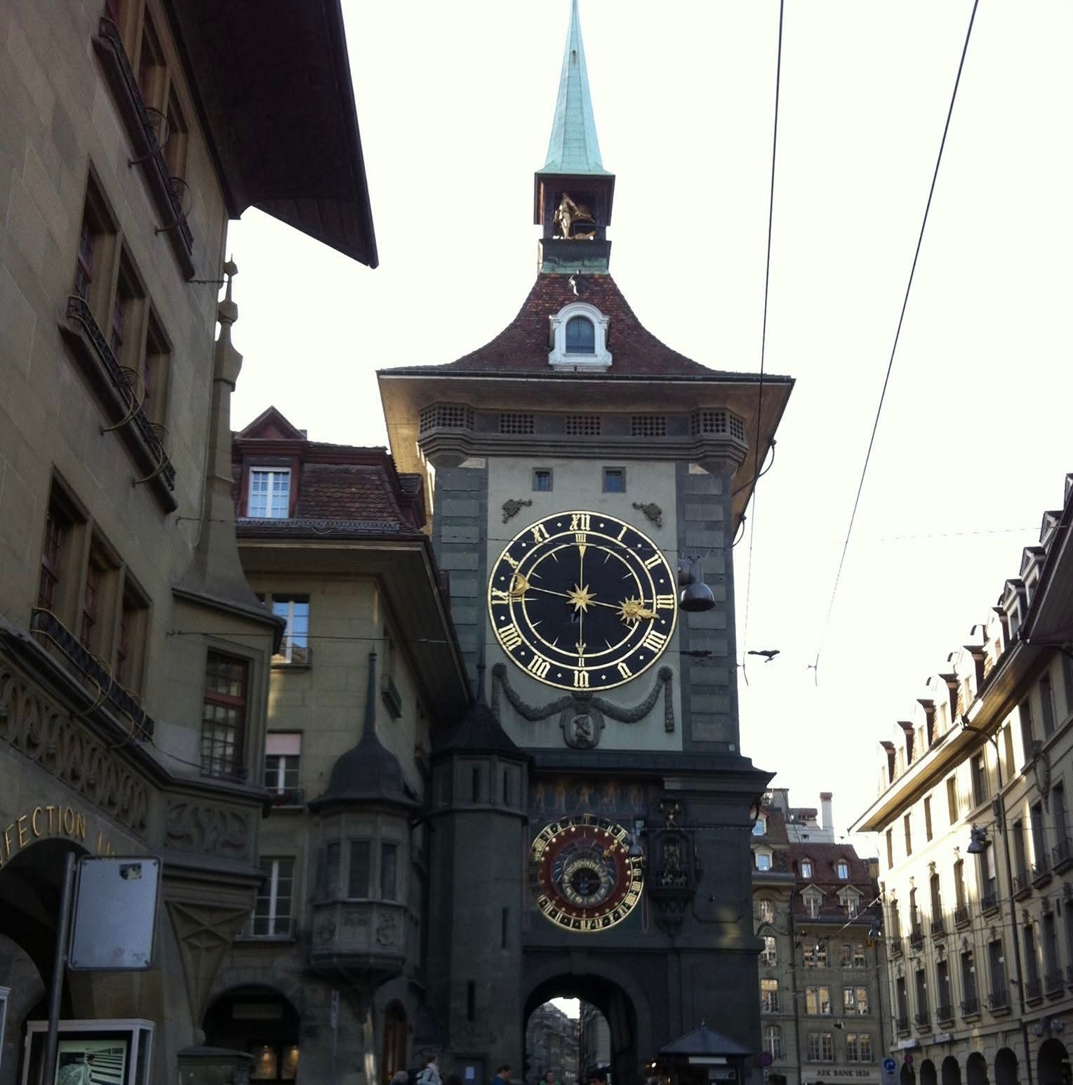 Historic clock tower with a large gold-and-black clock on a narrow street between old buildings