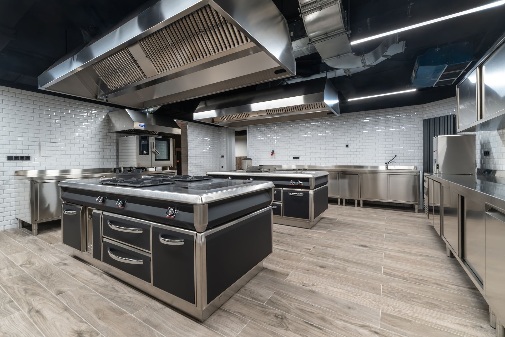 Professional commercial kitchen with stainless steel cooking ranges, large vent hoods, and light tile walls.